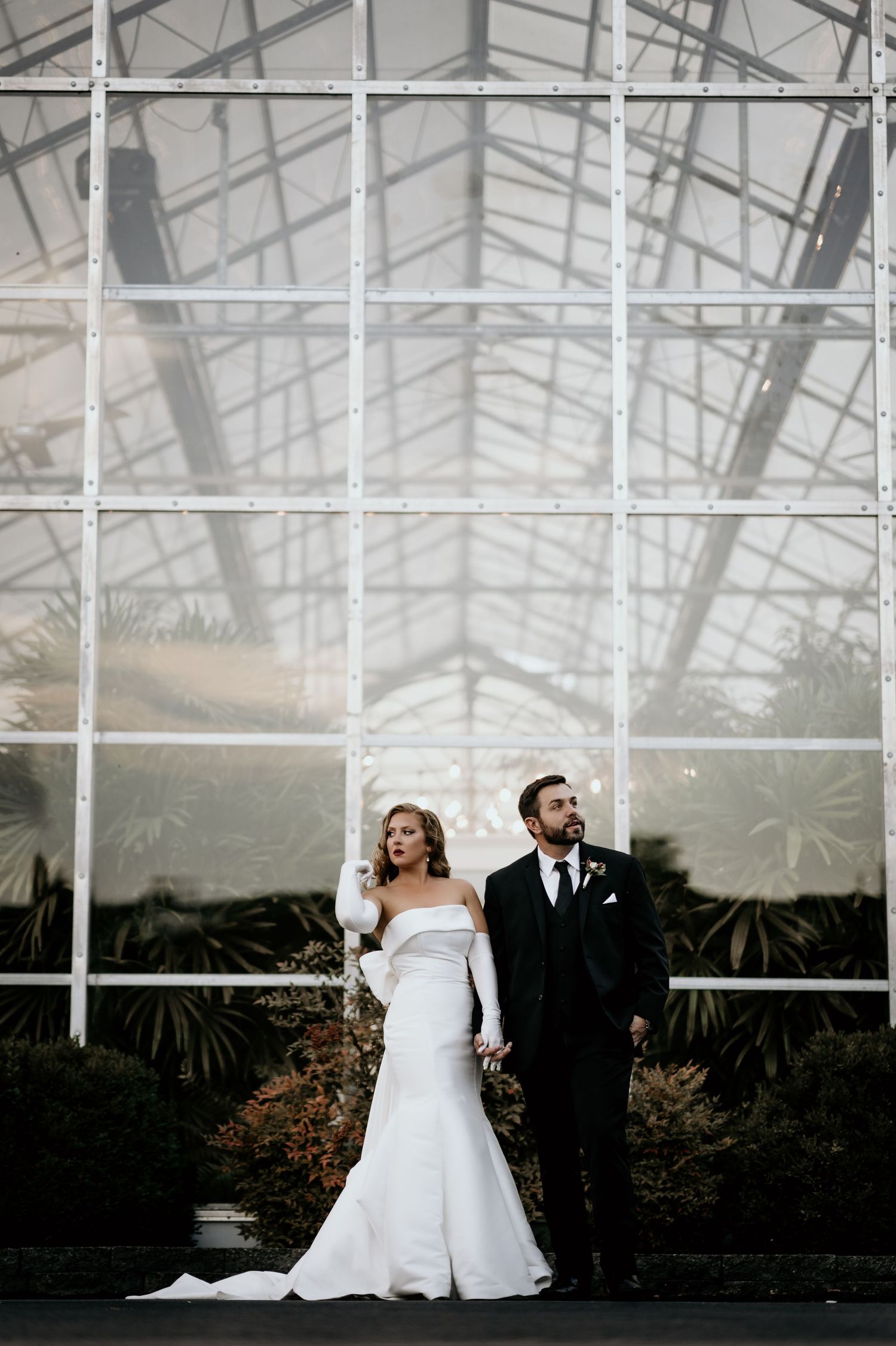 Elegant wedding couple poses in front of a grand greenhouse with dramatic glass architecture and lush greenery.