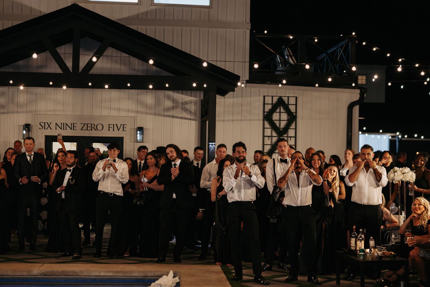 Wedding guests and groomsmen in formal attire gather on a dance floor under string lights at an indoor reception venue.