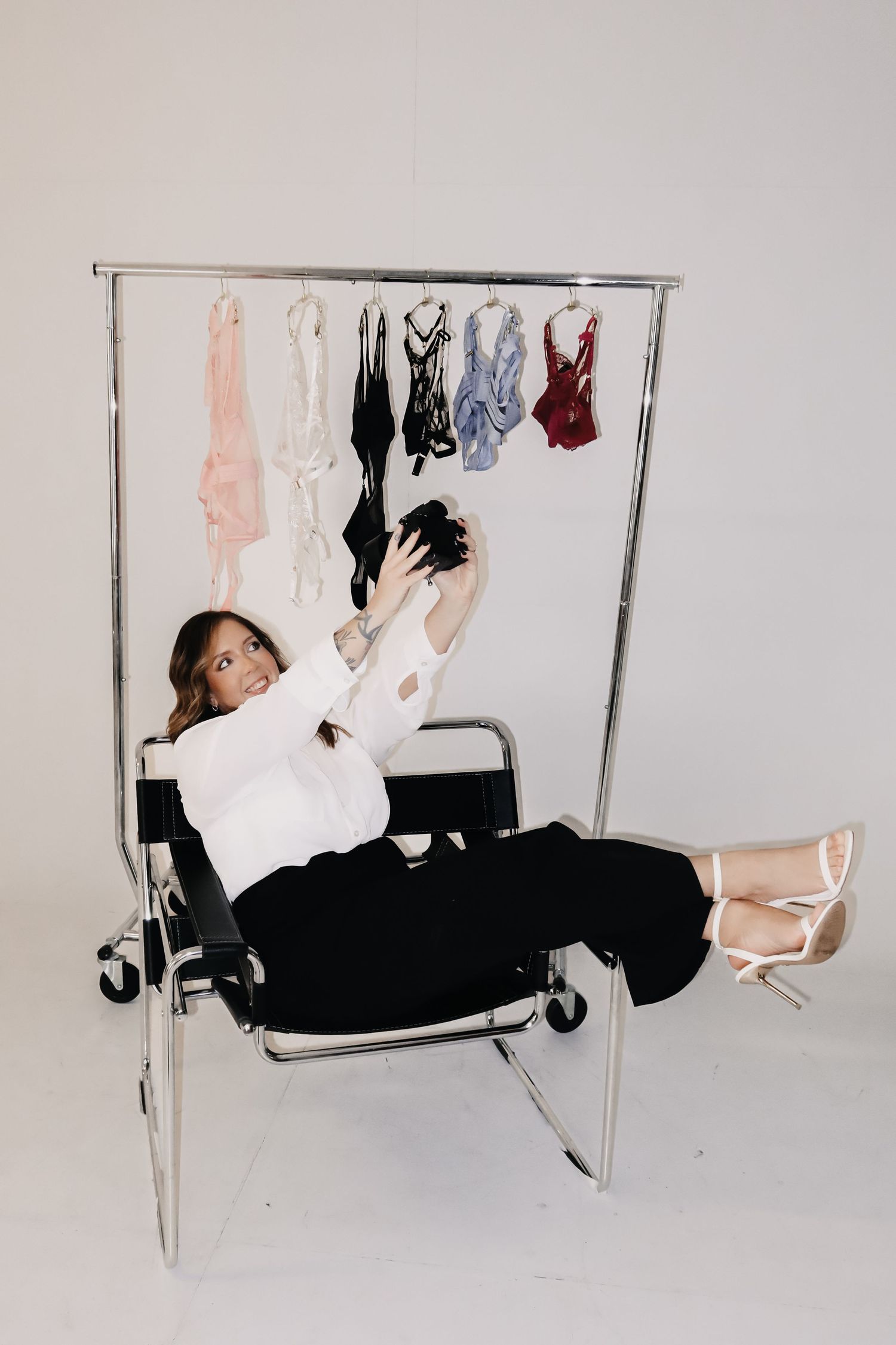 Woman in white blouse and black pants reclines on chair, taking selfie near clothing rack with lingerie.