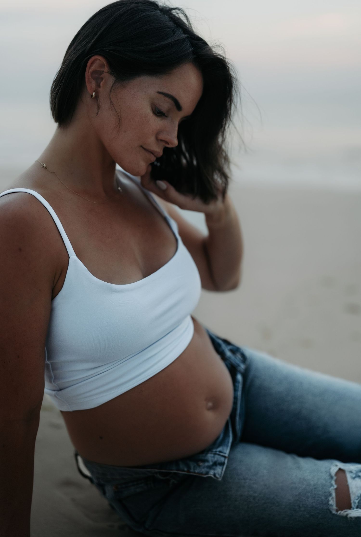 Pregnant woman in white crop top and jeans sitting on beach, cradling baby bump during golden hour.