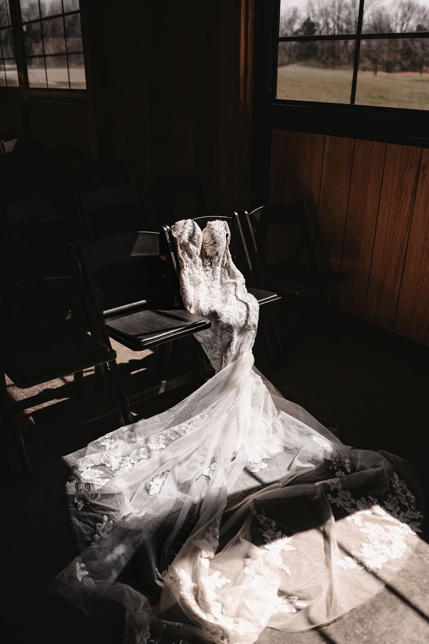 Elegant lace wedding dress with long train draped over wooden chairs in dramatic sunlight inside a rustic venue.