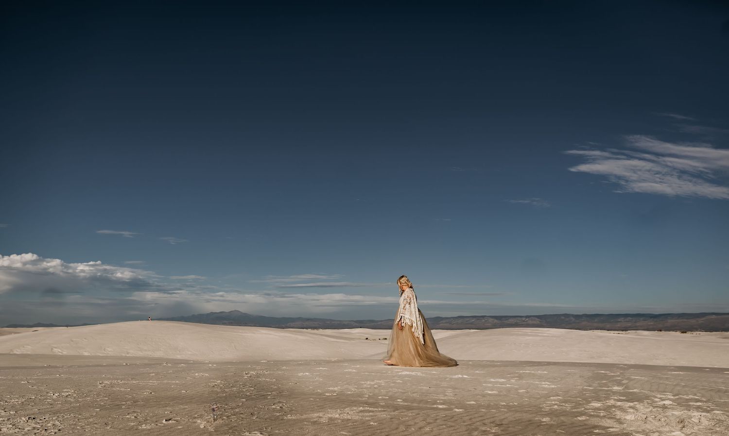A figure in a flowing champagne gown stands alone on a vast white salt flat under a dramatic blue desert sky.