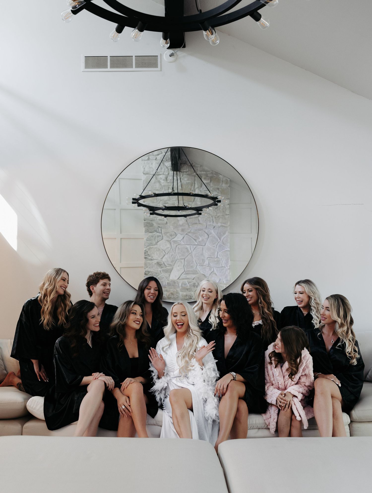 A group of women in black robes and one in white sit together on a couch under a round mirror and chandelier.