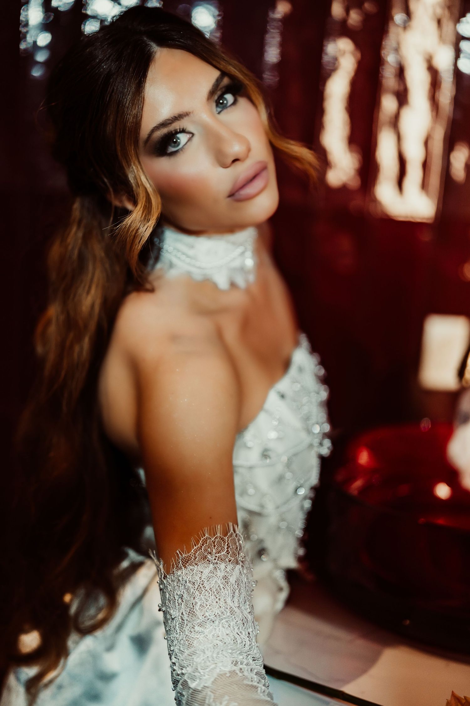 Elegant woman in white lace gown with choker necklace poses against a dark red bokeh background.