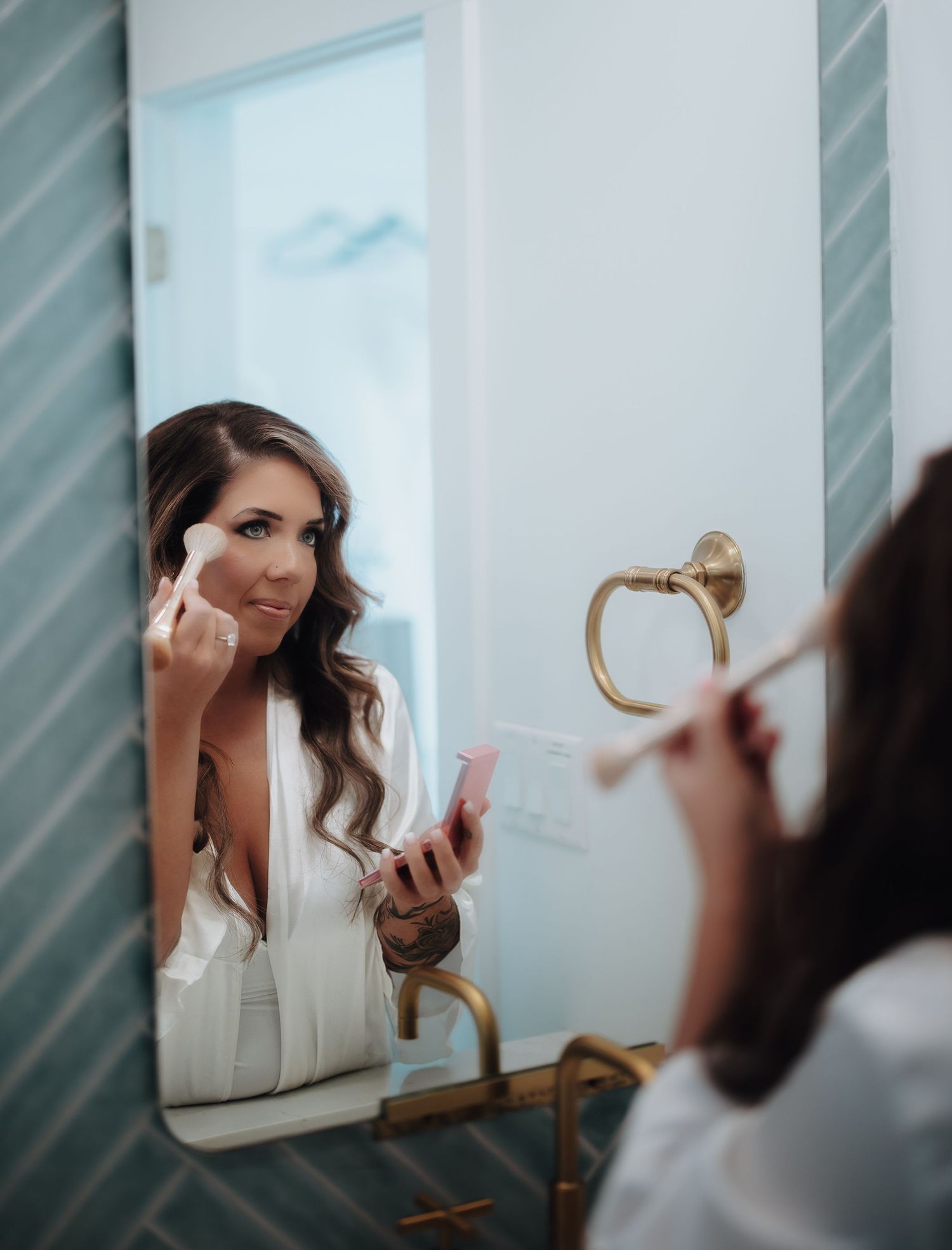A person with long dark hair is reflected in a mirror with a brass lamp mounted on the wall.
