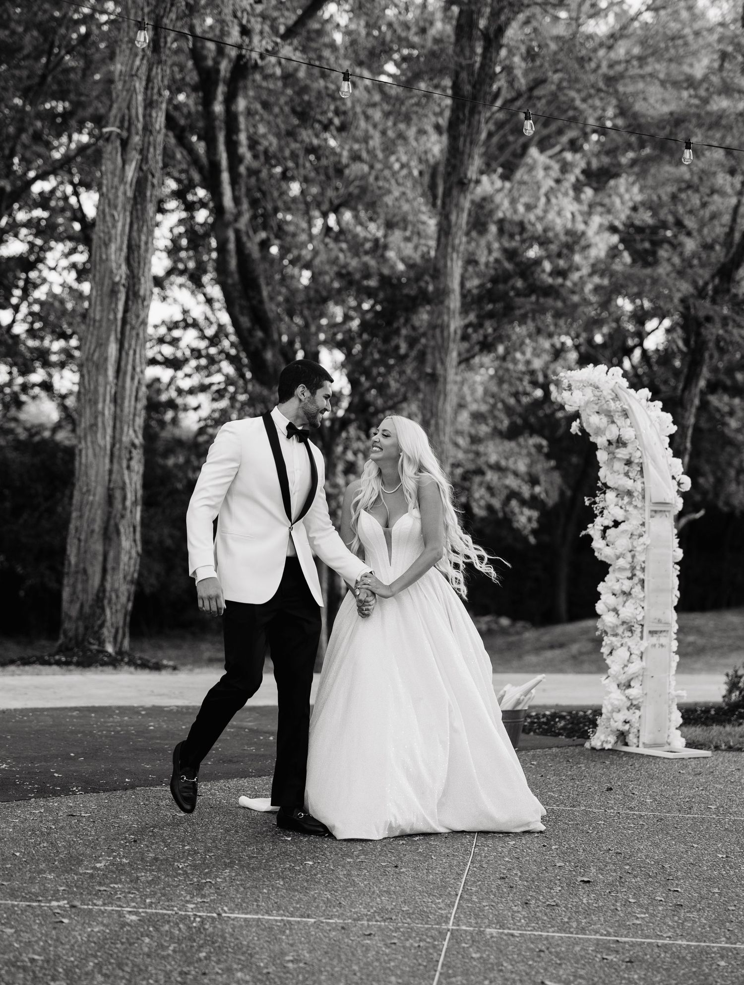 Black and white wedding photo with couple walking together outdoors near a decorative floral arch.