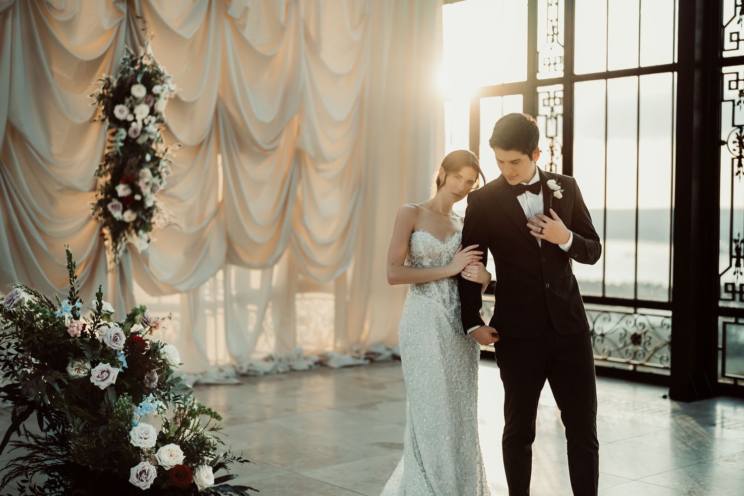 Elegant wedding couple in a sunlit venue with floral arrangements, groom in black suit and bride in white gown.