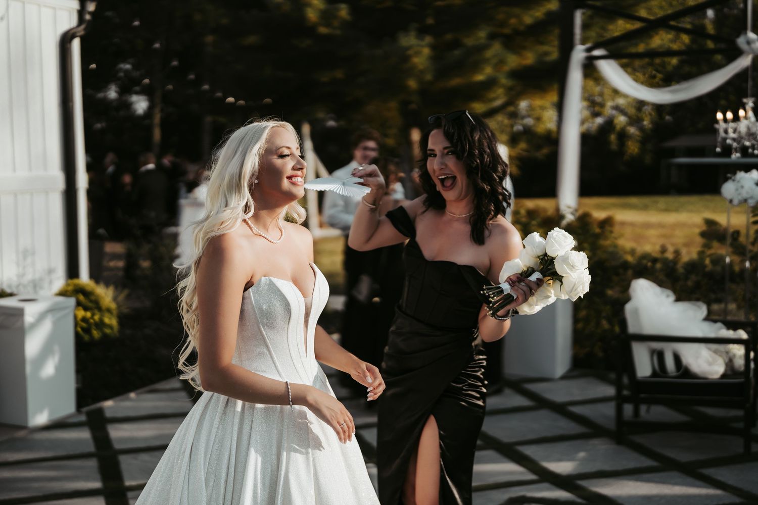 Bride in white strapless gown walks with bridesmaid in black dress holding bouquet at outdoor wedding.