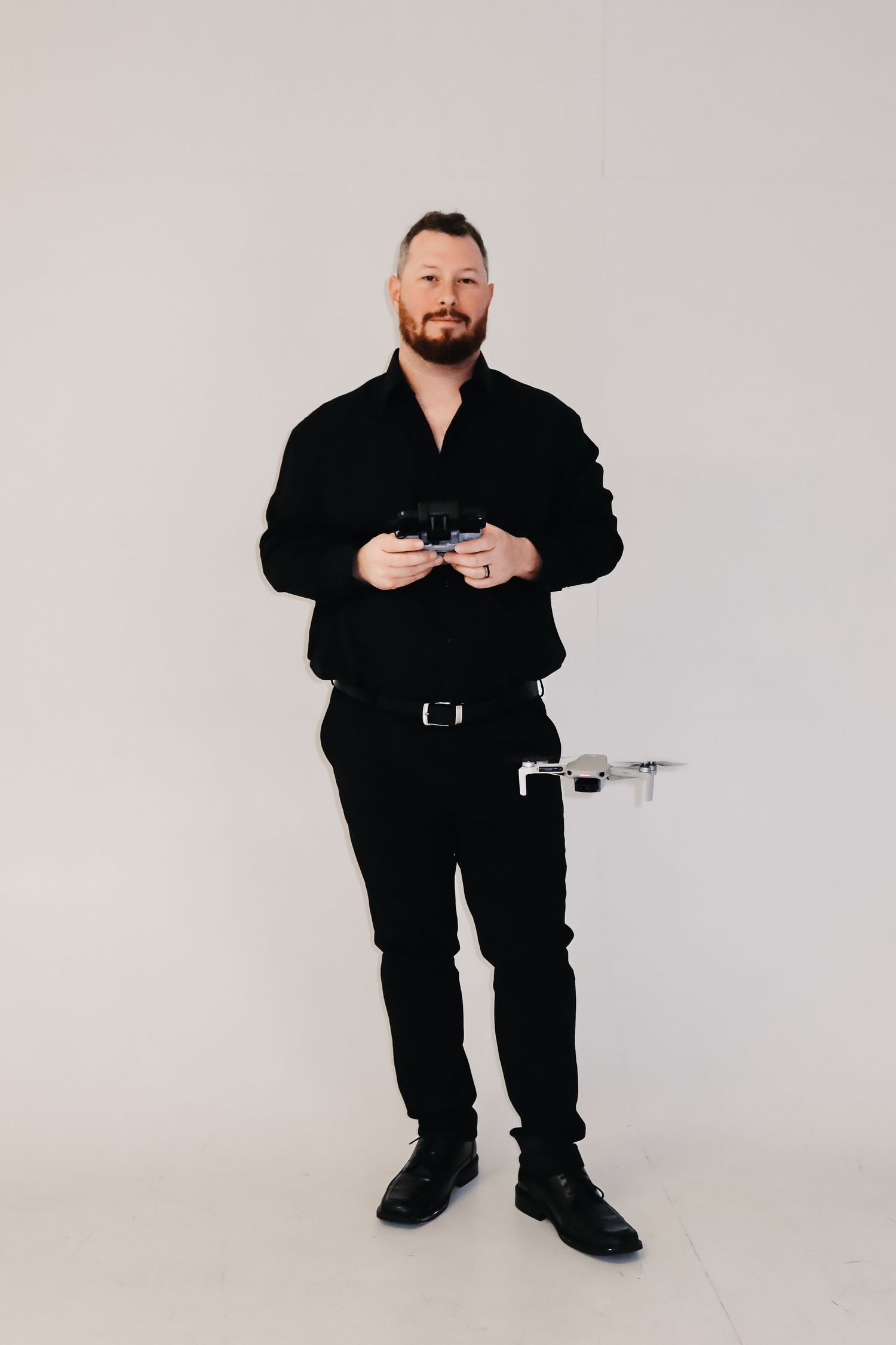 Bearded man in black outfit holding a drone controller with a small drone, standing against a white background.