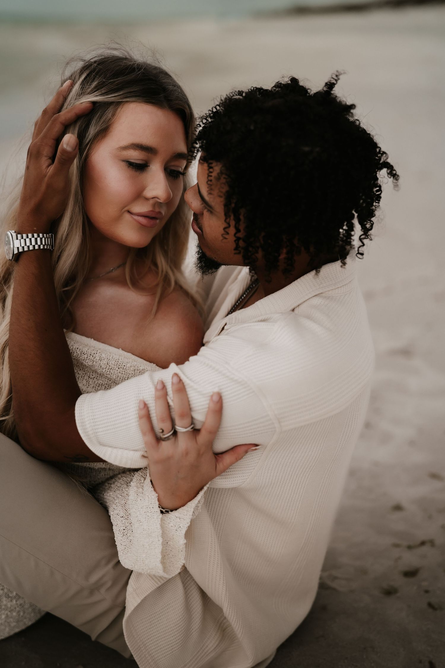 Interracial couple sharing a romantic embrace on the beach, both dressed in white outfits.