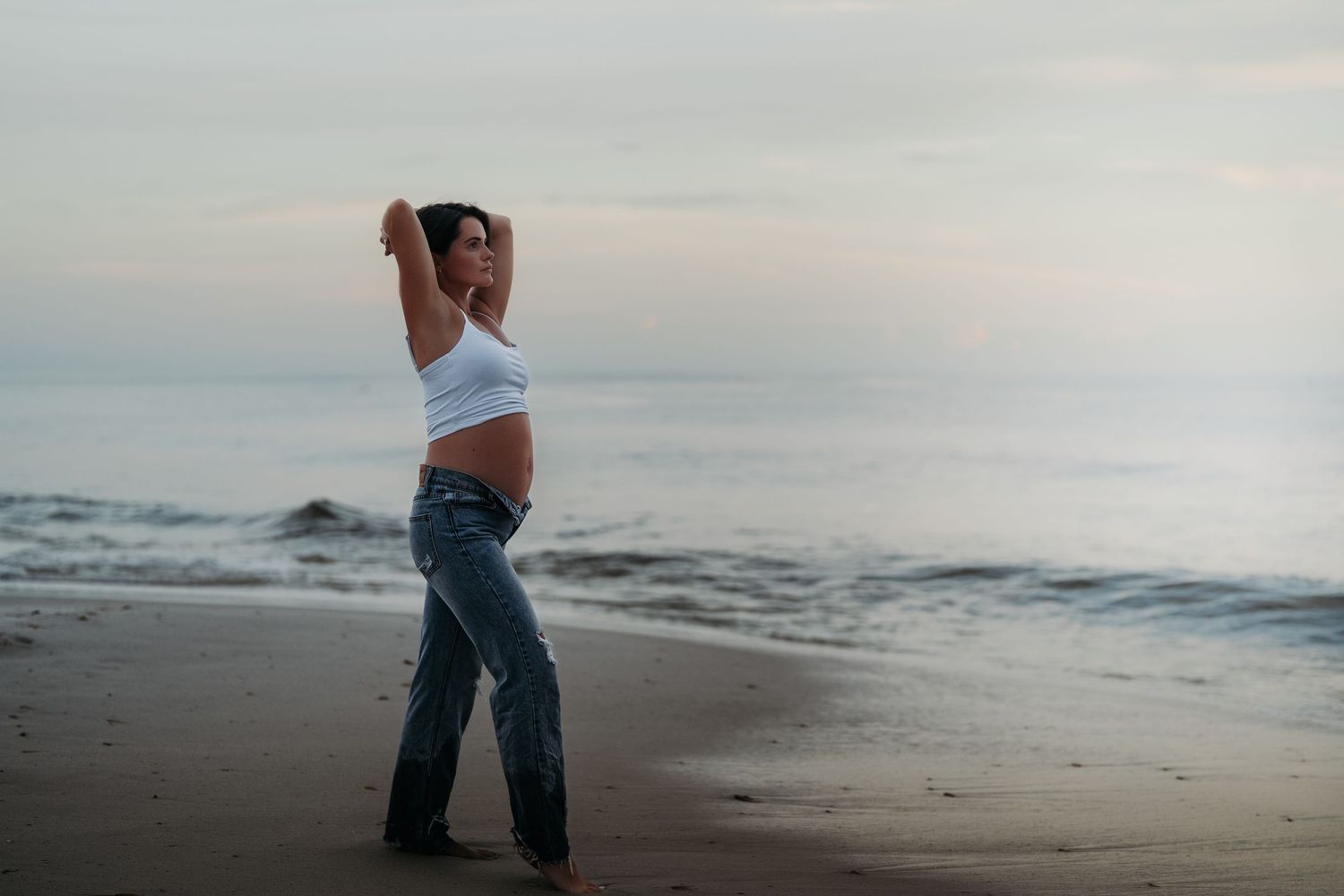 Pregnant woman in white crop top and jeans posing on a serene beach at dusk with ocean waves behind her.