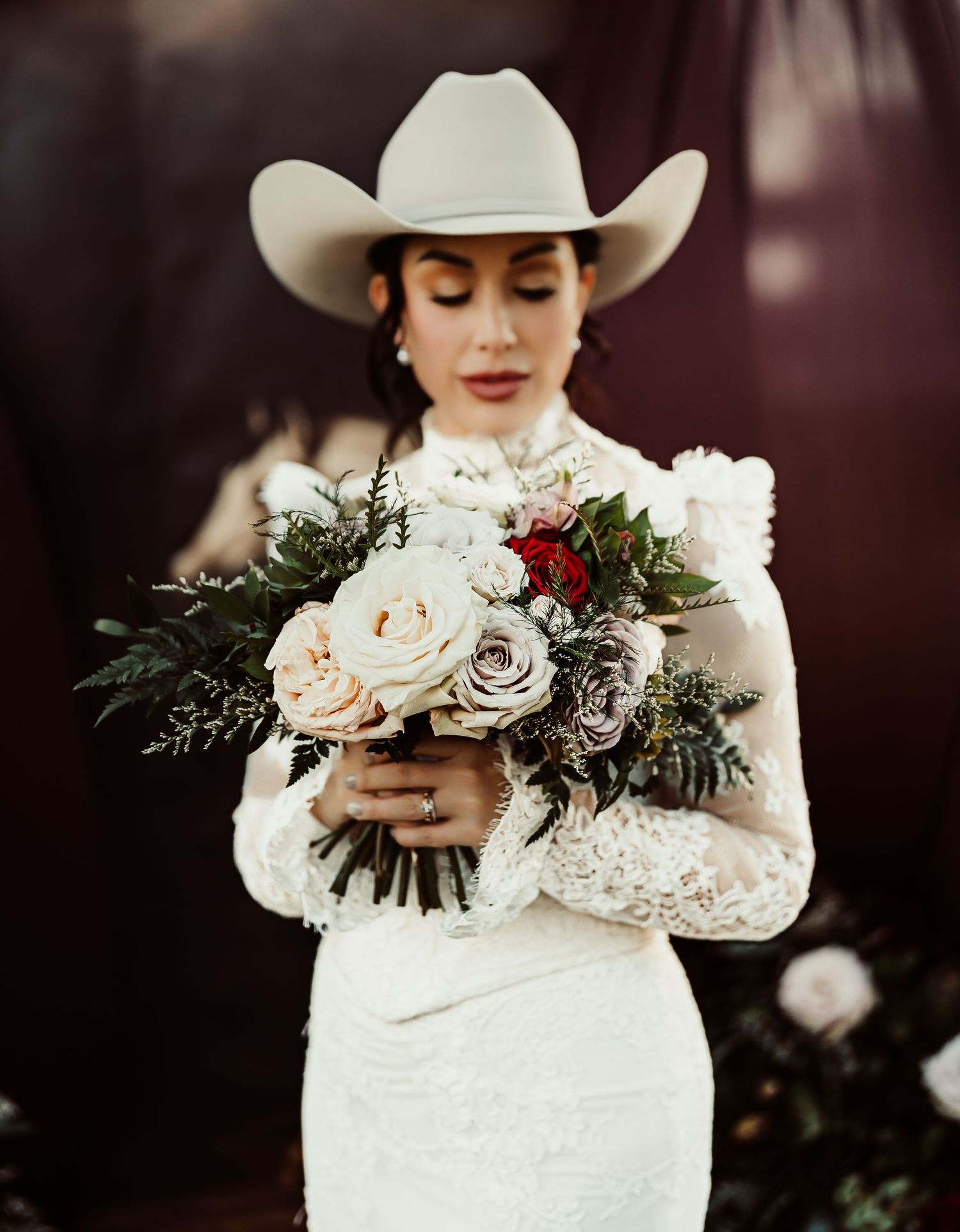 Bride in white lace gown and cowboy hat holding a rustic bouquet of roses and greenery at a western-themed wedding.