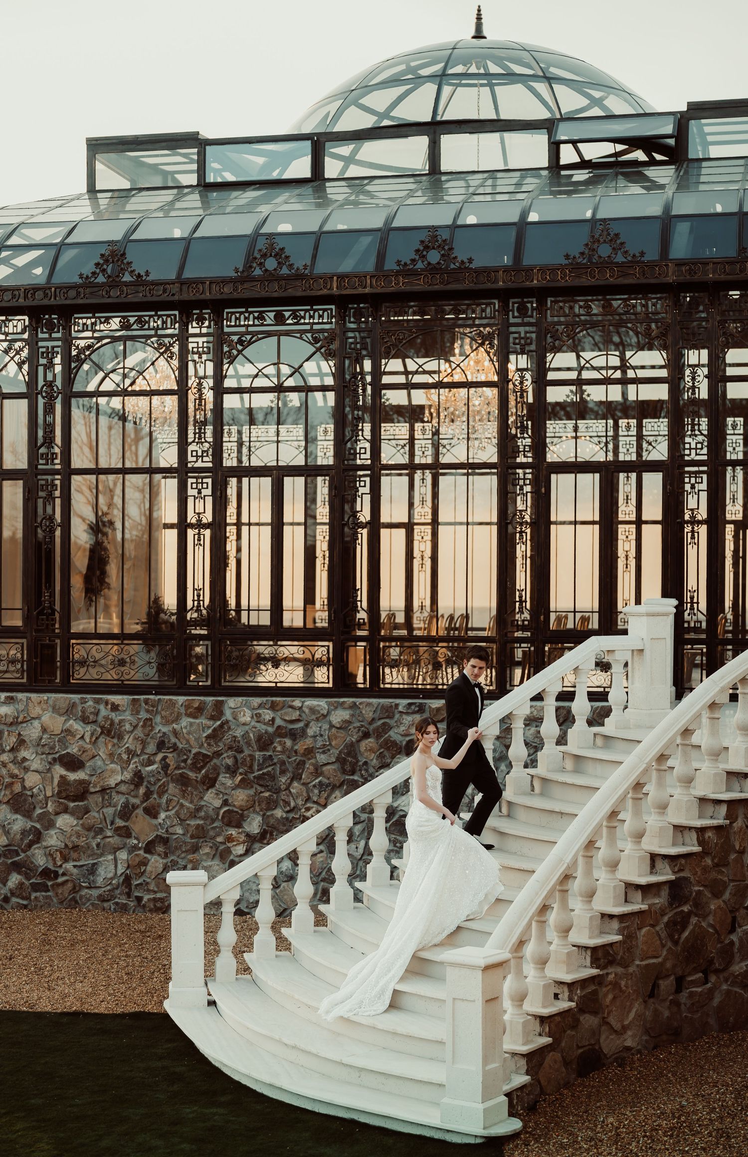 Bride and groom pose on elegant white staircase before ornate Victorian greenhouse at golden hour wedding.