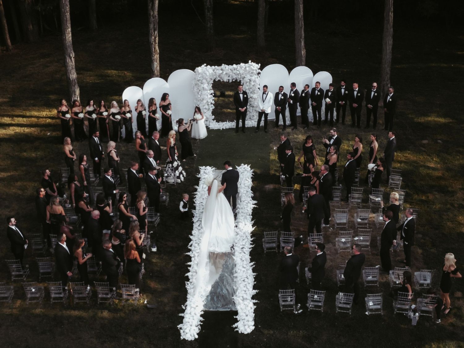 Aerial view of an outdoor wedding ceremony with white floral arch and guests seated in a curved formation in the woods.