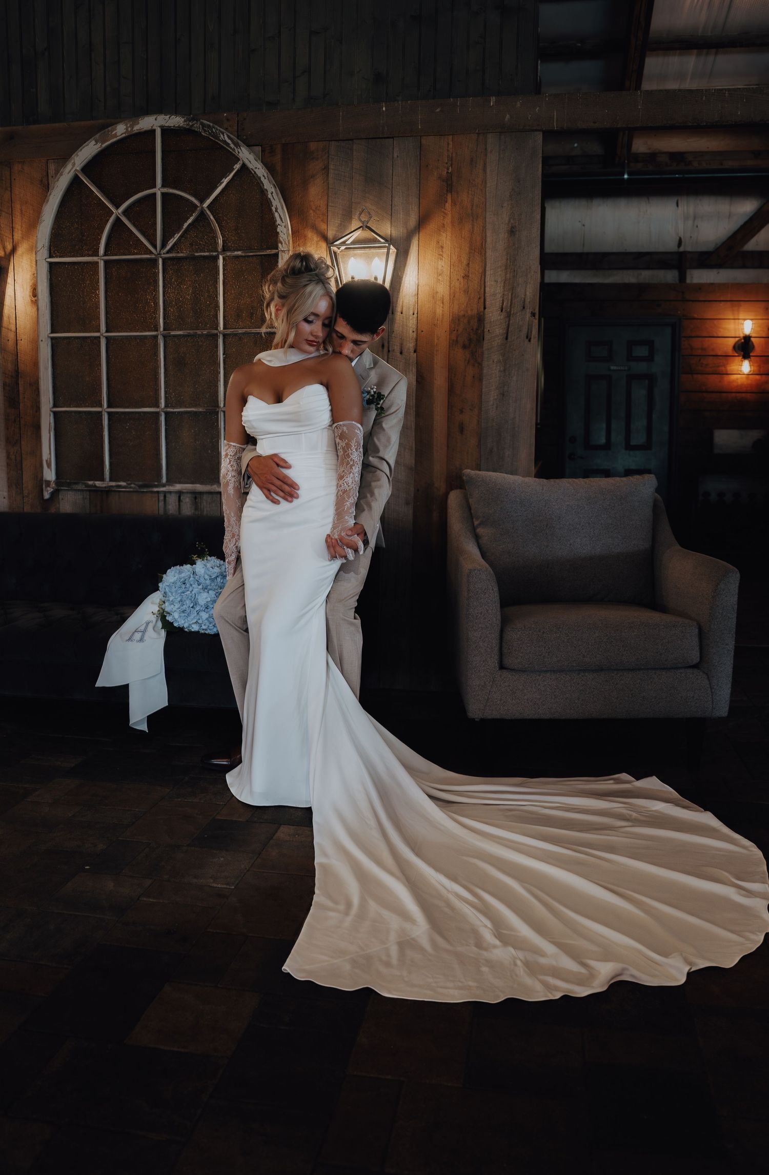 Bride in elegant white strapless gown with train stands with groom in rustic barn venue with wooden walls.