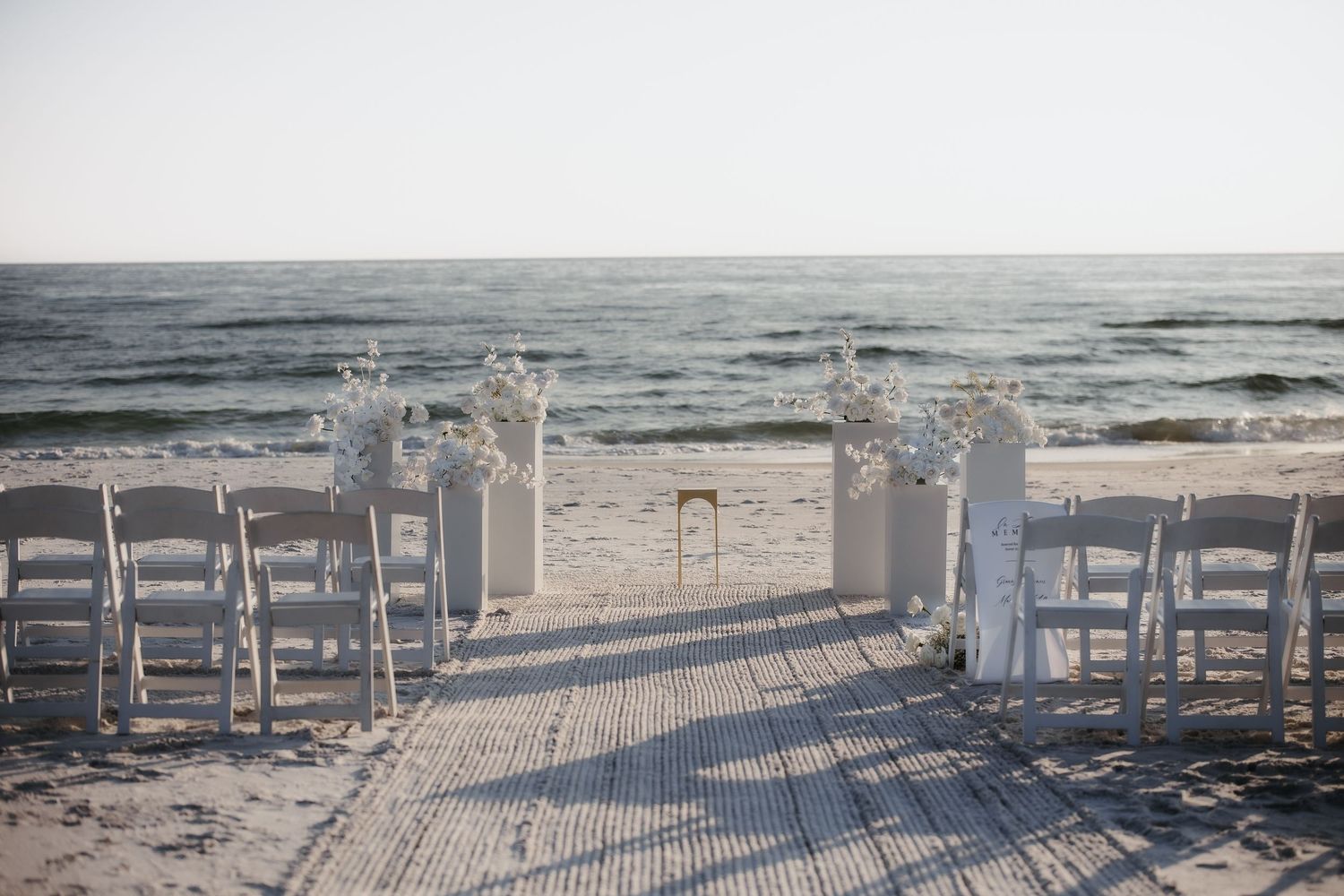 White wooden chairs arranged in rows on a sandy beach for a seaside wedding ceremony at sunset.