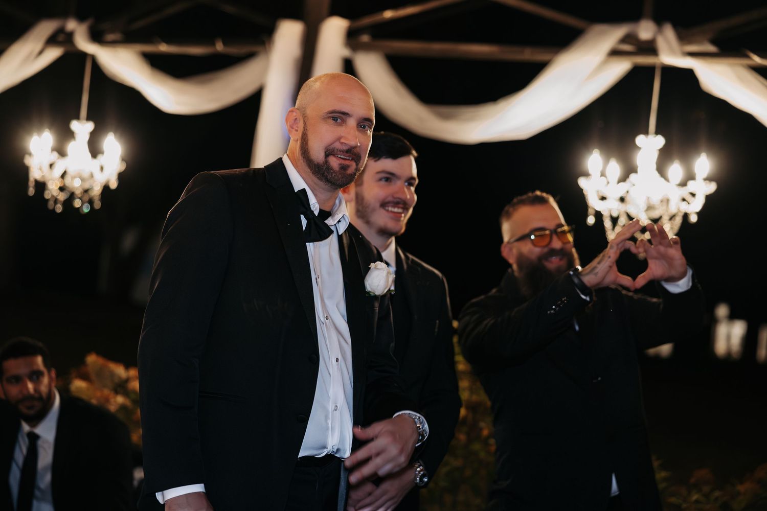 Three men in tuxedos posing at a formal event with elegant chandeliers in the background.