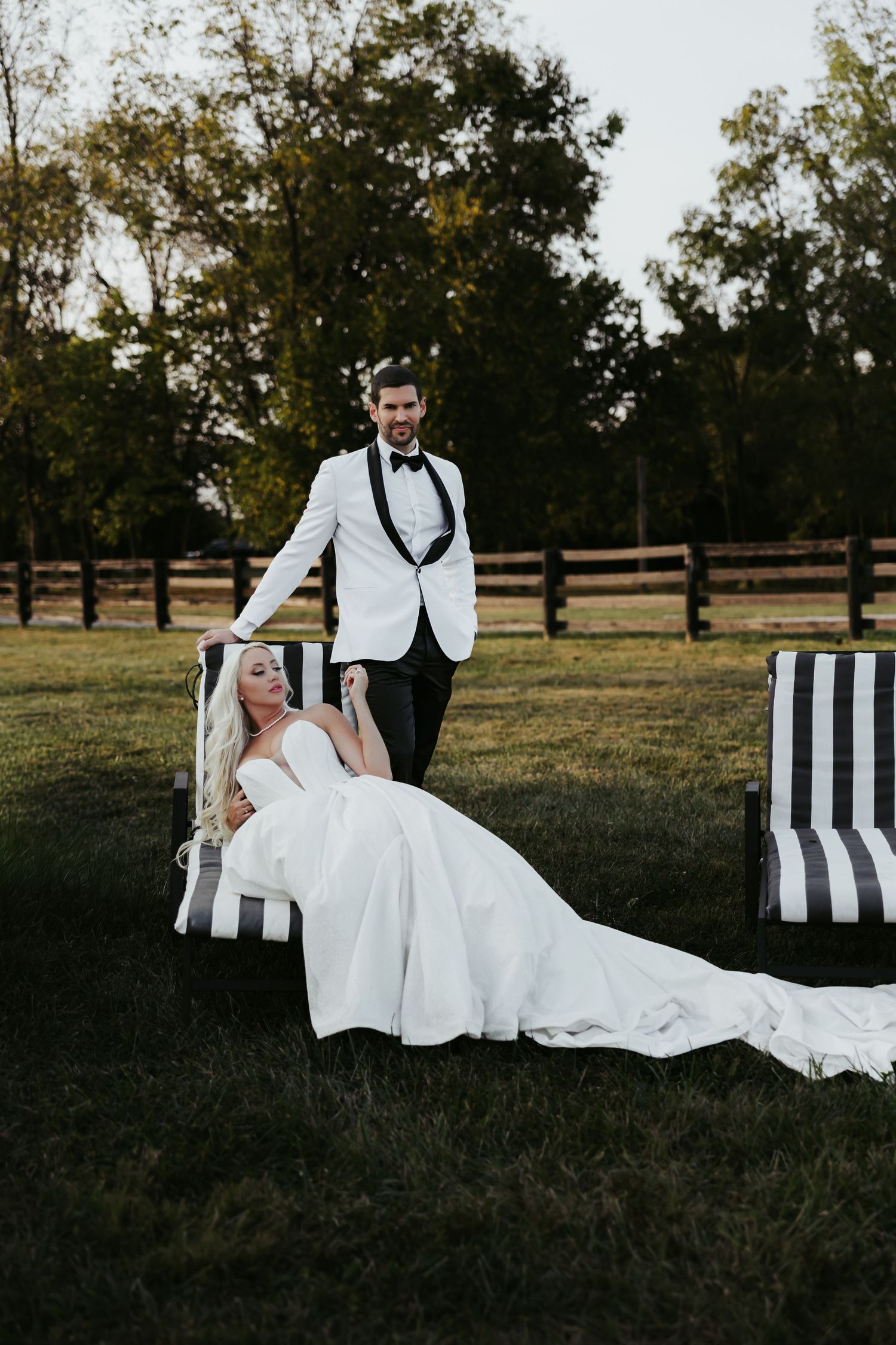 Bride in white gown reclines on striped chair outdoors while groom in white tuxedo stands beside her on green lawn.