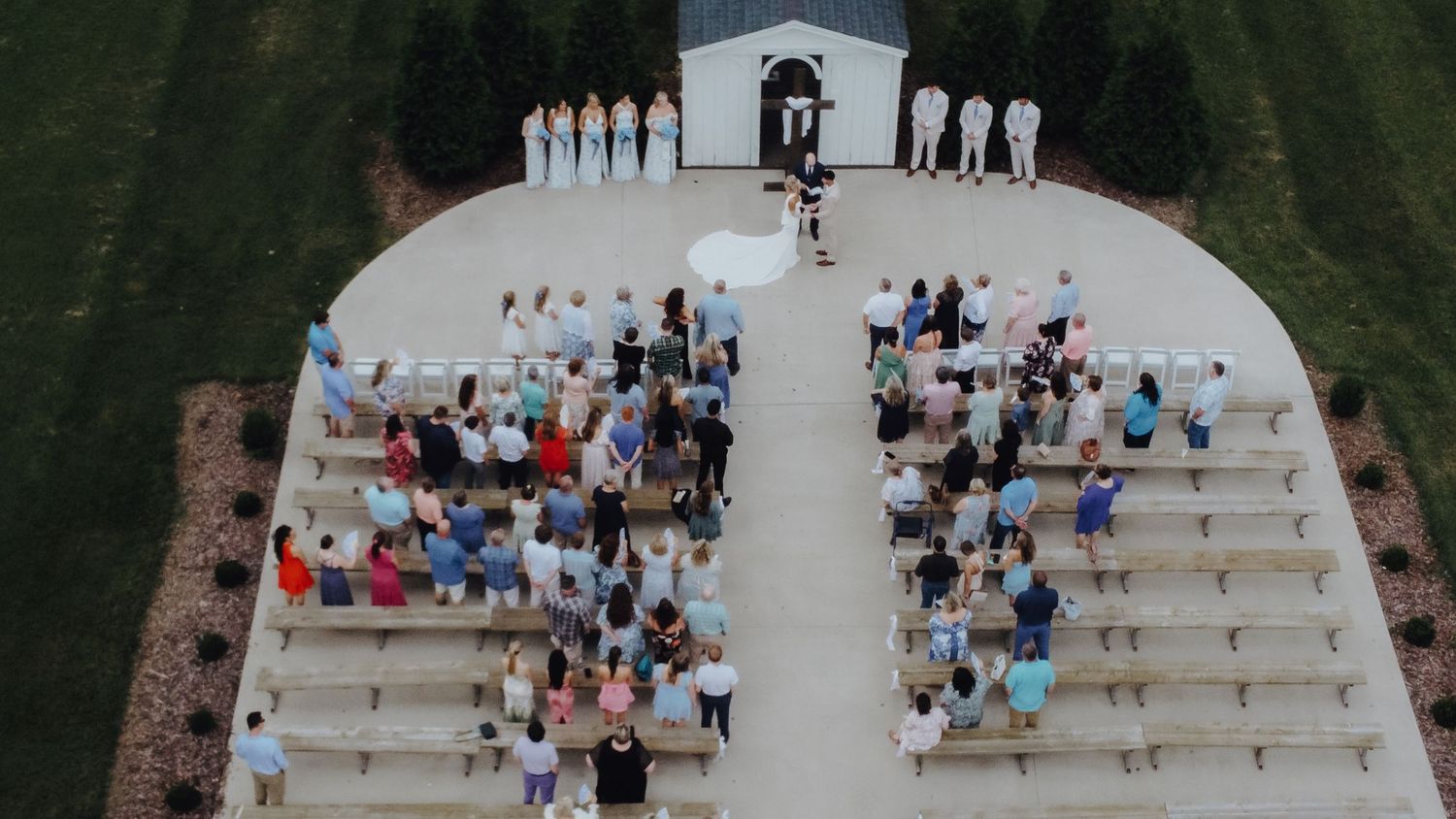 Aerial view of an outdoor wedding ceremony with guests seated in curved rows before a white chapel.