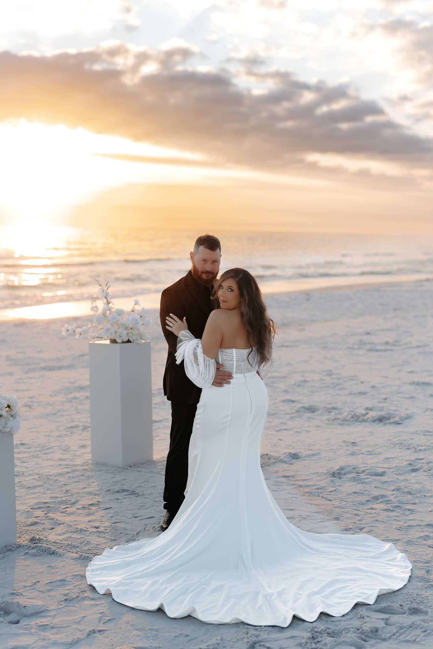 A romantic beach wedding scene at sunset with a couple on white sand as waves crash in the background.