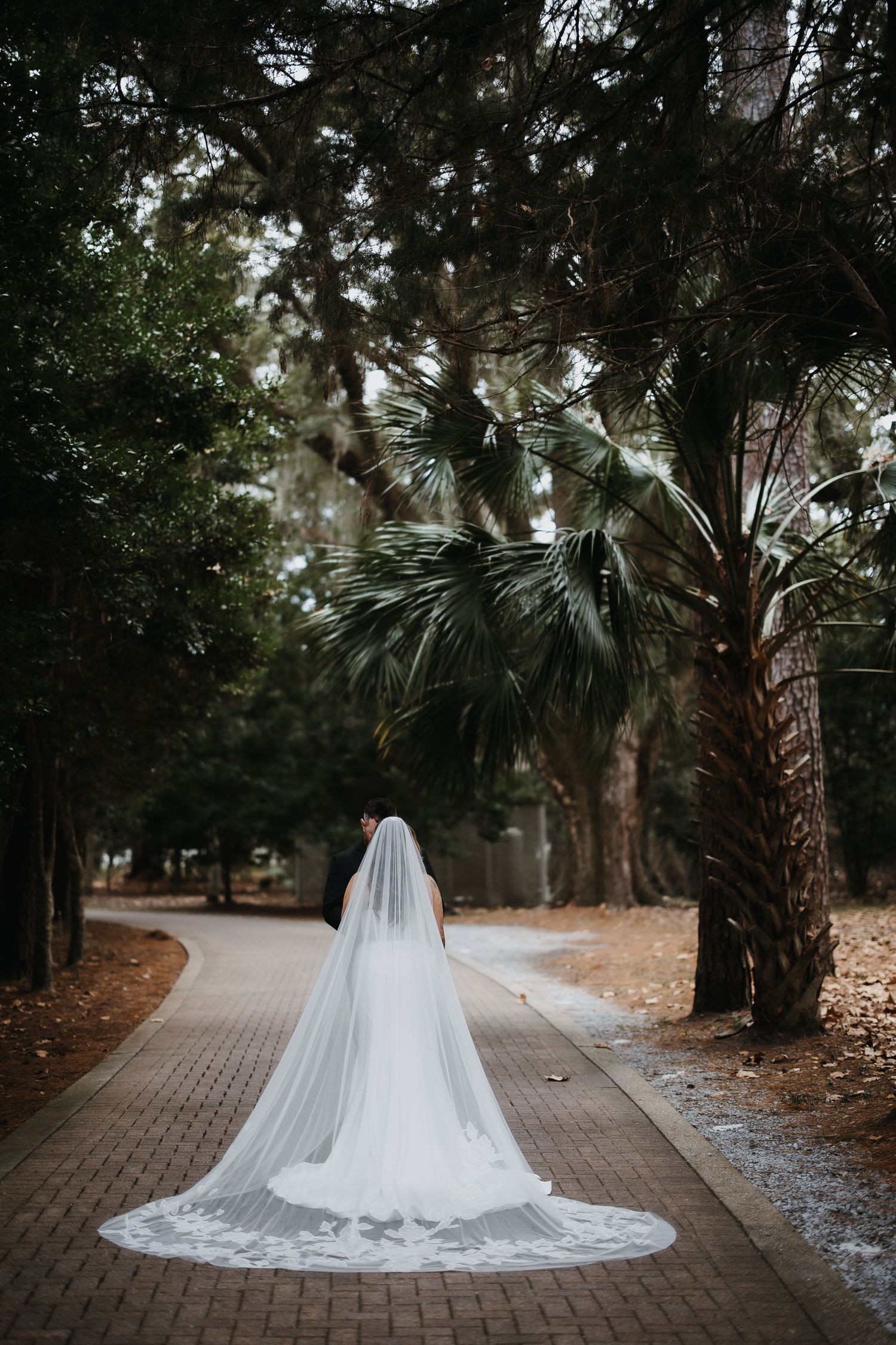 A flowing white wedding veil trails along a brick pathway surrounded by palm trees and greenery in a moody garden setting.