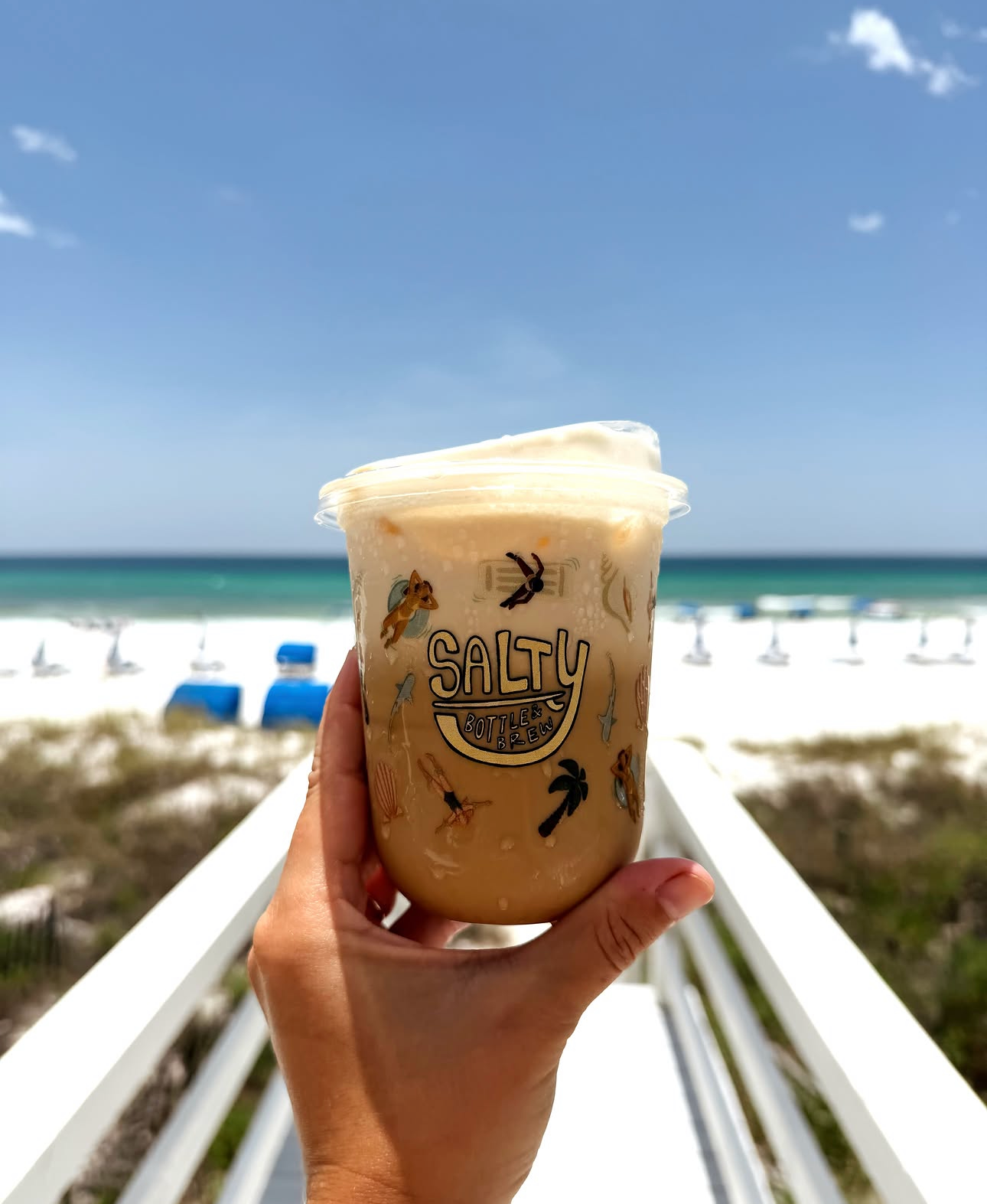 Hand holding a Salty branded iced coffee drink on a sunny beach boardwalk with turquoise ocean in the background.