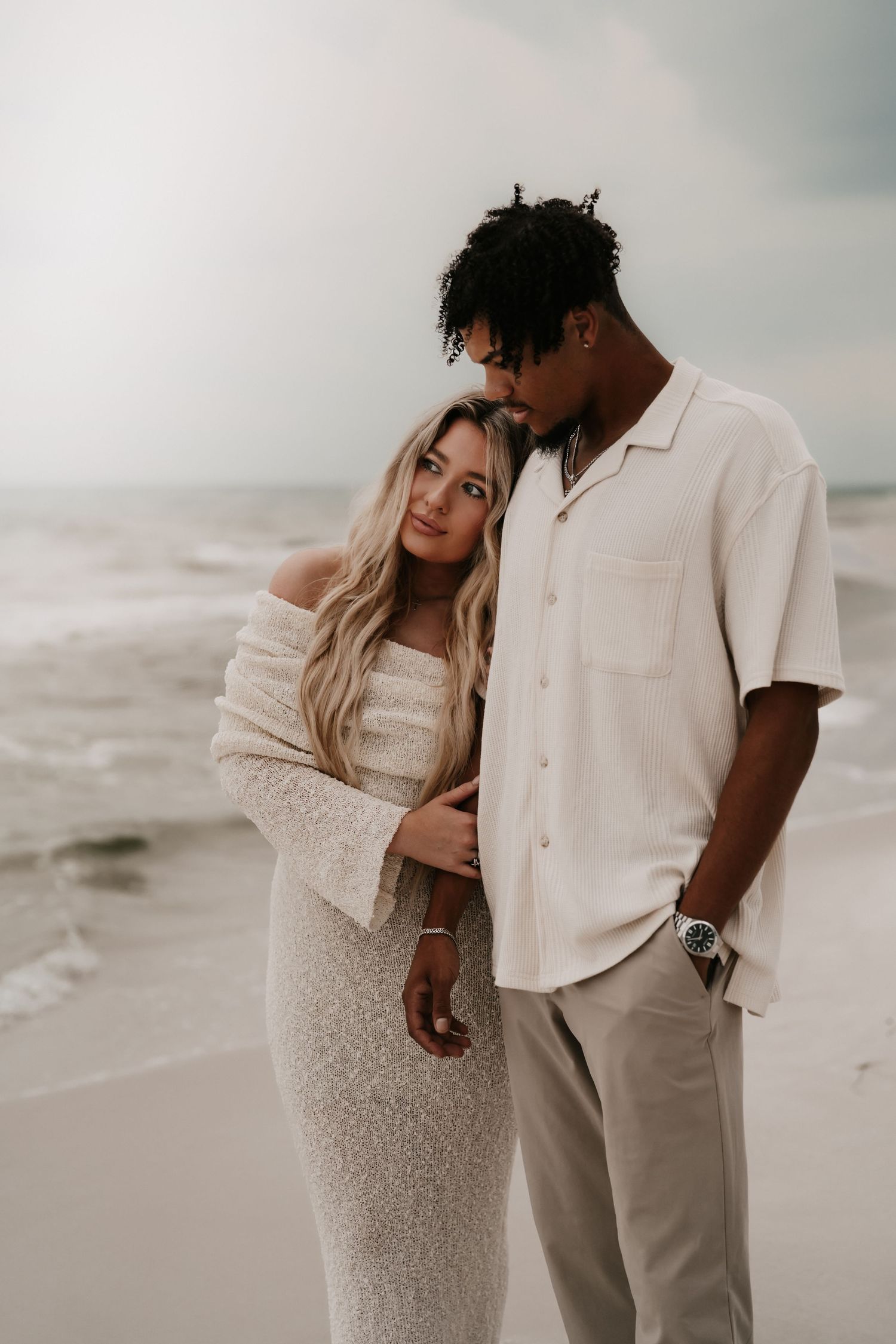 A romantic couple embraces on a cloudy beach, wearing coordinated white and cream colored outfits.
