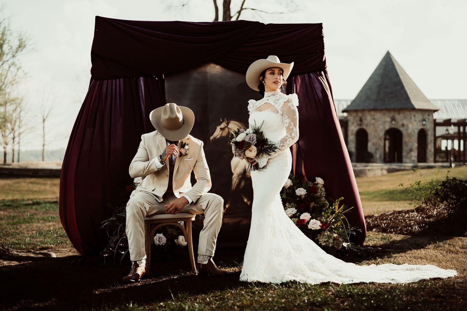 Bride in white gown and cowboy hat poses with groom under burgundy drape at rustic outdoor wedding venue.