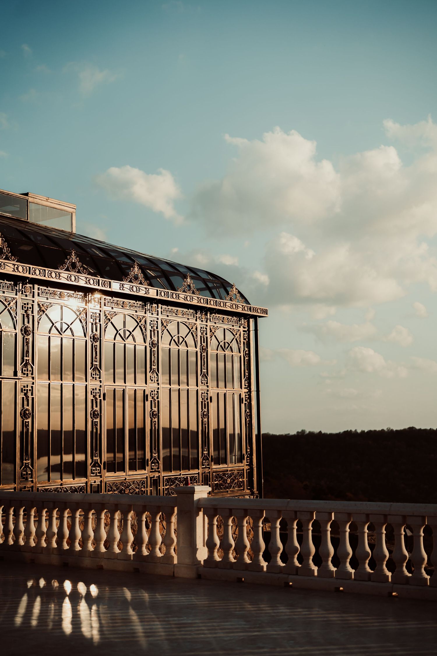 Ornate Victorian glass greenhouse with golden ironwork on a terrace balustrade at sunset against a blue sky.