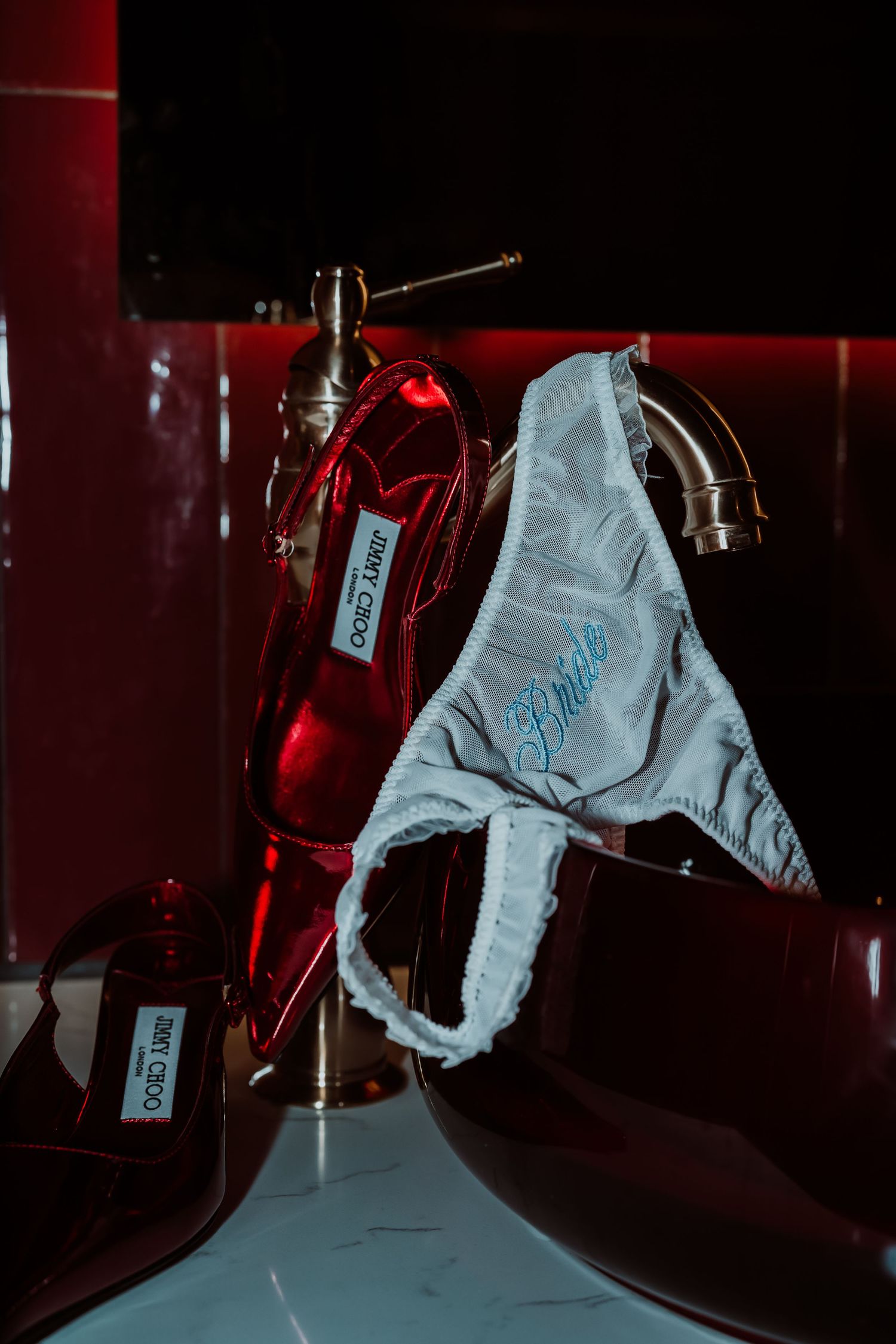 White satin bride thong underwear draped over red Jimmy Choo heels on a sink in moody red lighting.