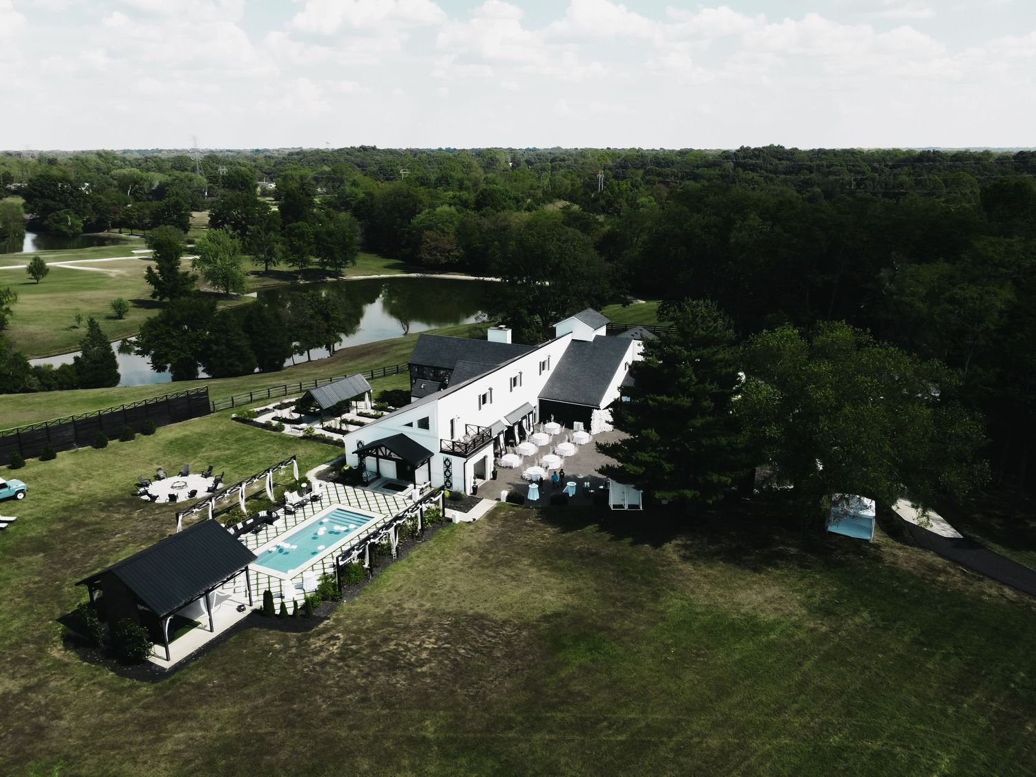 Aerial panoramic view of a white event venue with tents and swimming pool surrounded by lush green landscape.