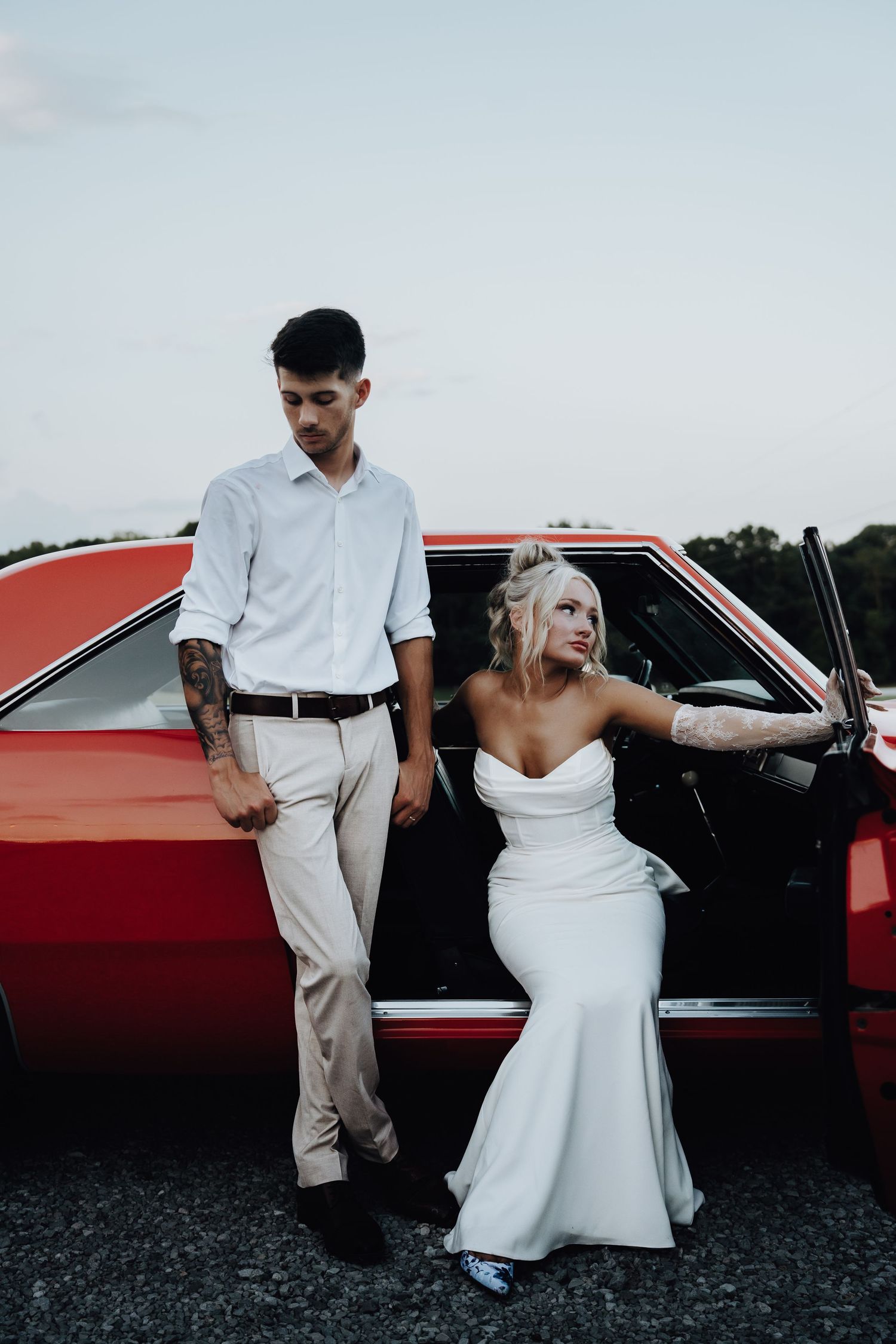 Bride in white mermaid gown and groom in white shirt pose beside a classic red car outdoors.