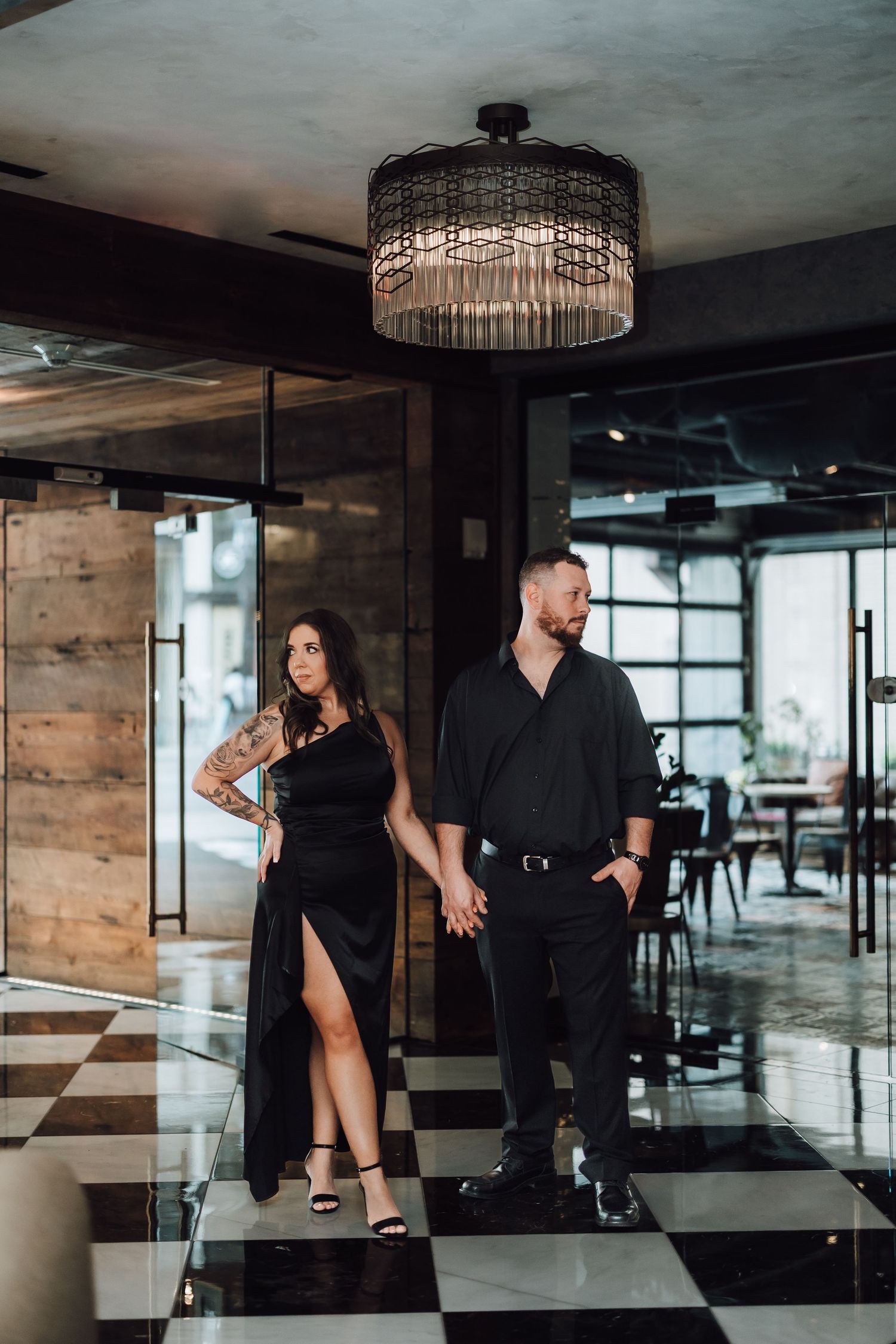 A couple in elegant black attire poses on a checkered floor under a crystal chandelier in a modern restaurant interior.