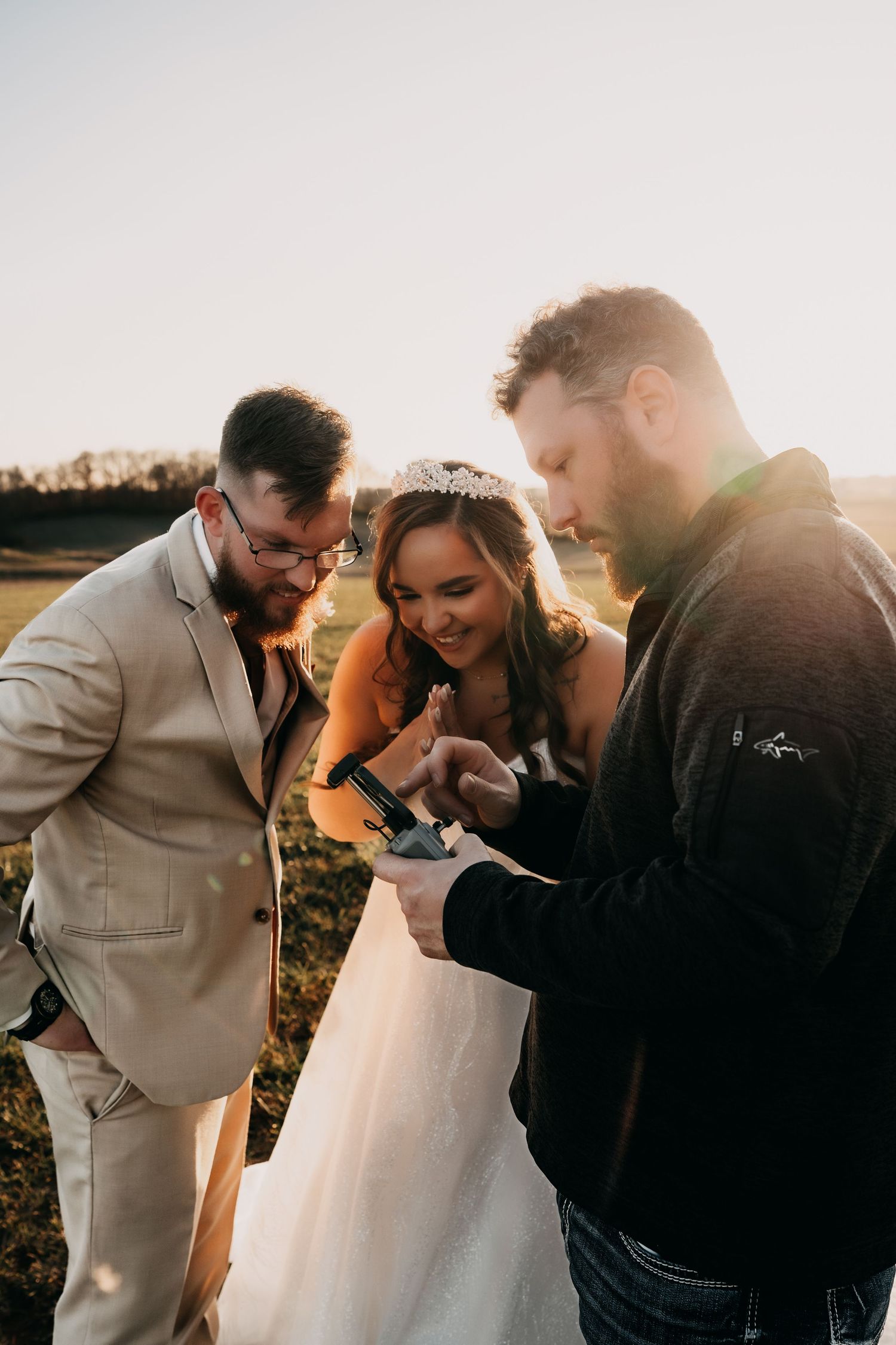 Three people gathered outdoors at sunset looking at something together during a wedding ceremony.