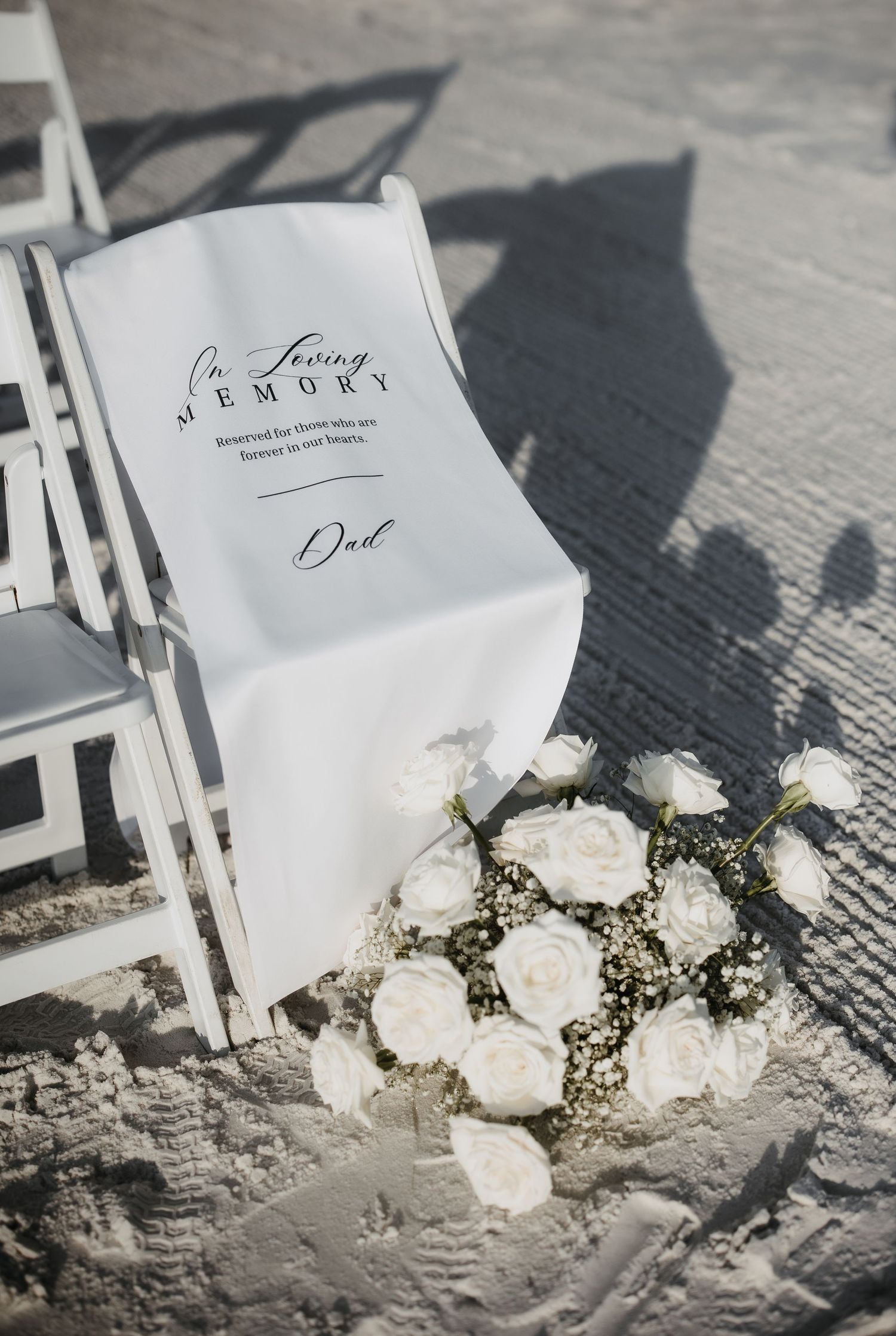 White wedding chair with a custom sign and elegant white rose bouquet on sandy beach during outdoor ceremony setup.