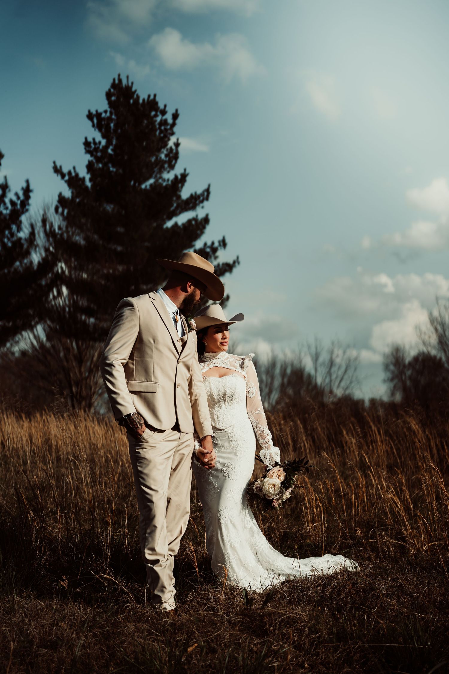 A bride and groom in cowboy hats hold hands in a golden field with a pine tree and blue sky backdrop.
