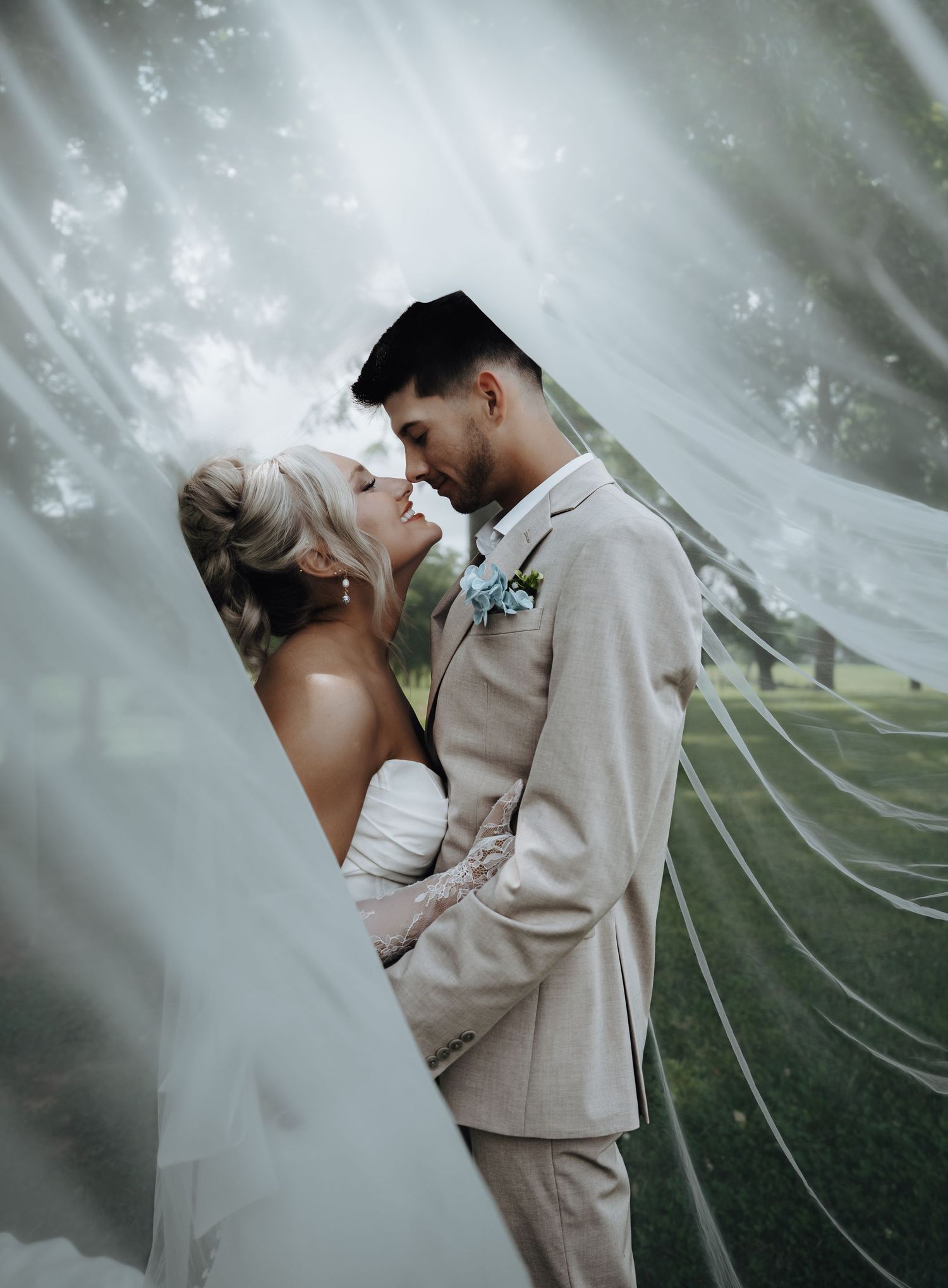 Bride and groom share a romantic almost-kiss beneath a flowing wedding veil outdoors on their wedding day.