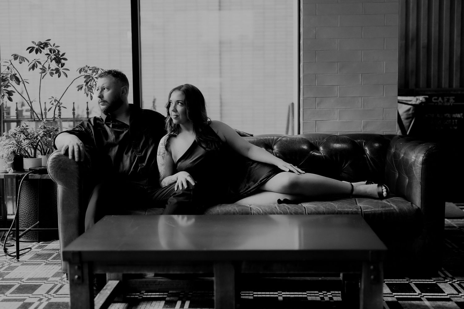 Couple posing on leather sofa in black and white photo, woman reclining elegantly while man sits beside her.