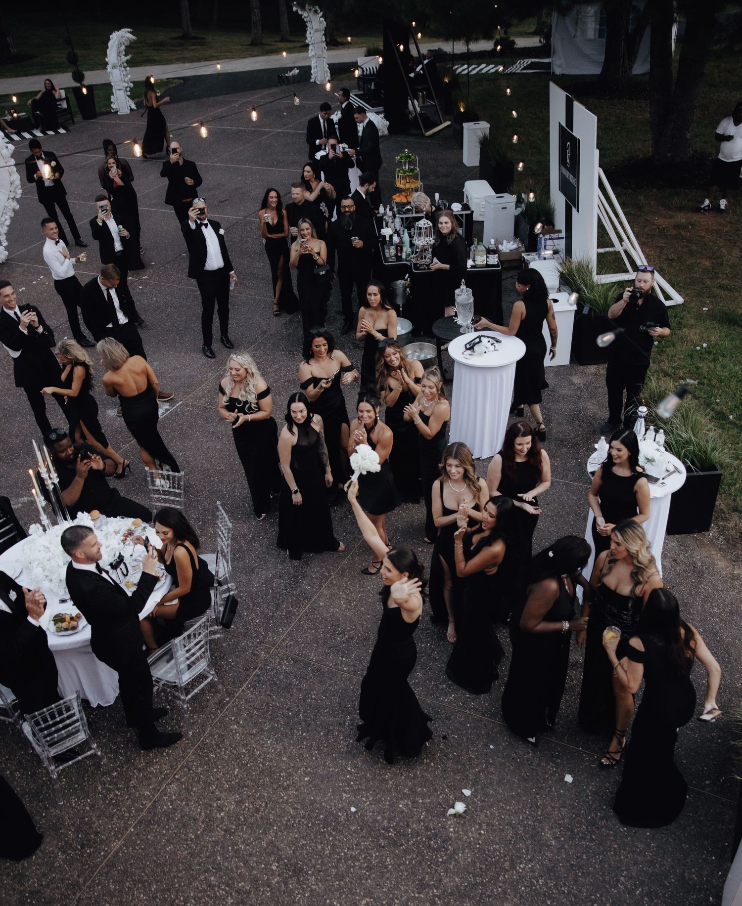 Aerial view of elegant black-tie wedding reception outdoors, guests in black formal attire mingling on patio with cake table.