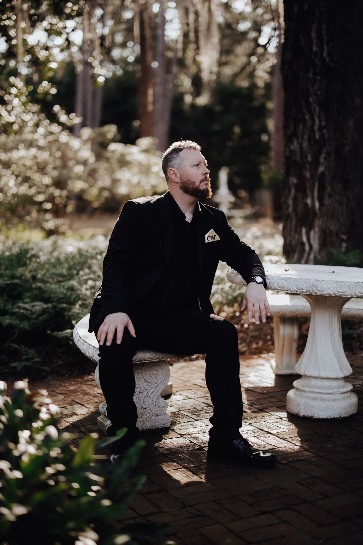 A person in black clothing sits contemplatively on a stone bench in a wooded outdoor setting.