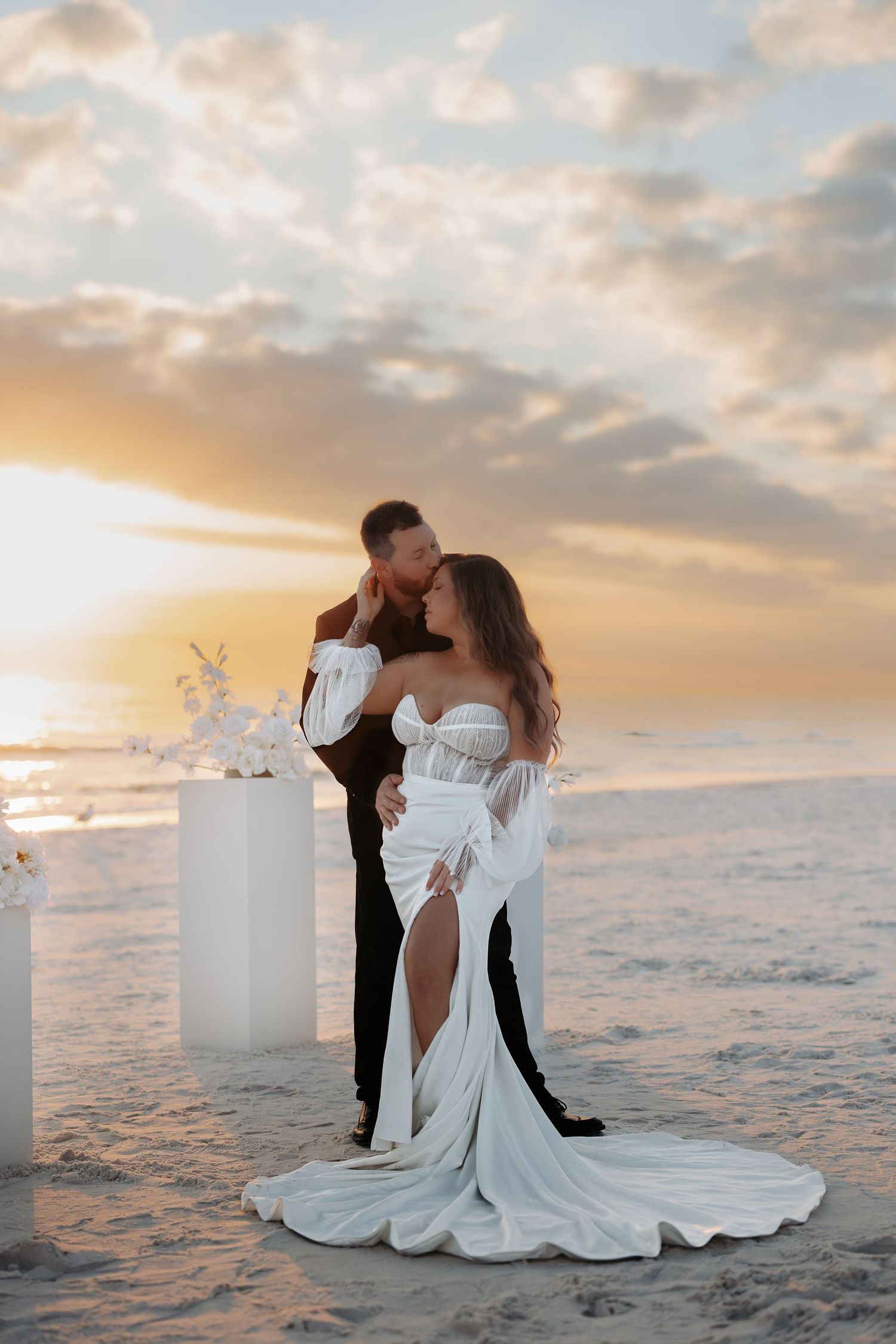 A romantic sunset beach wedding photoshoot with a couple posing on white furniture against an ocean backdrop.
