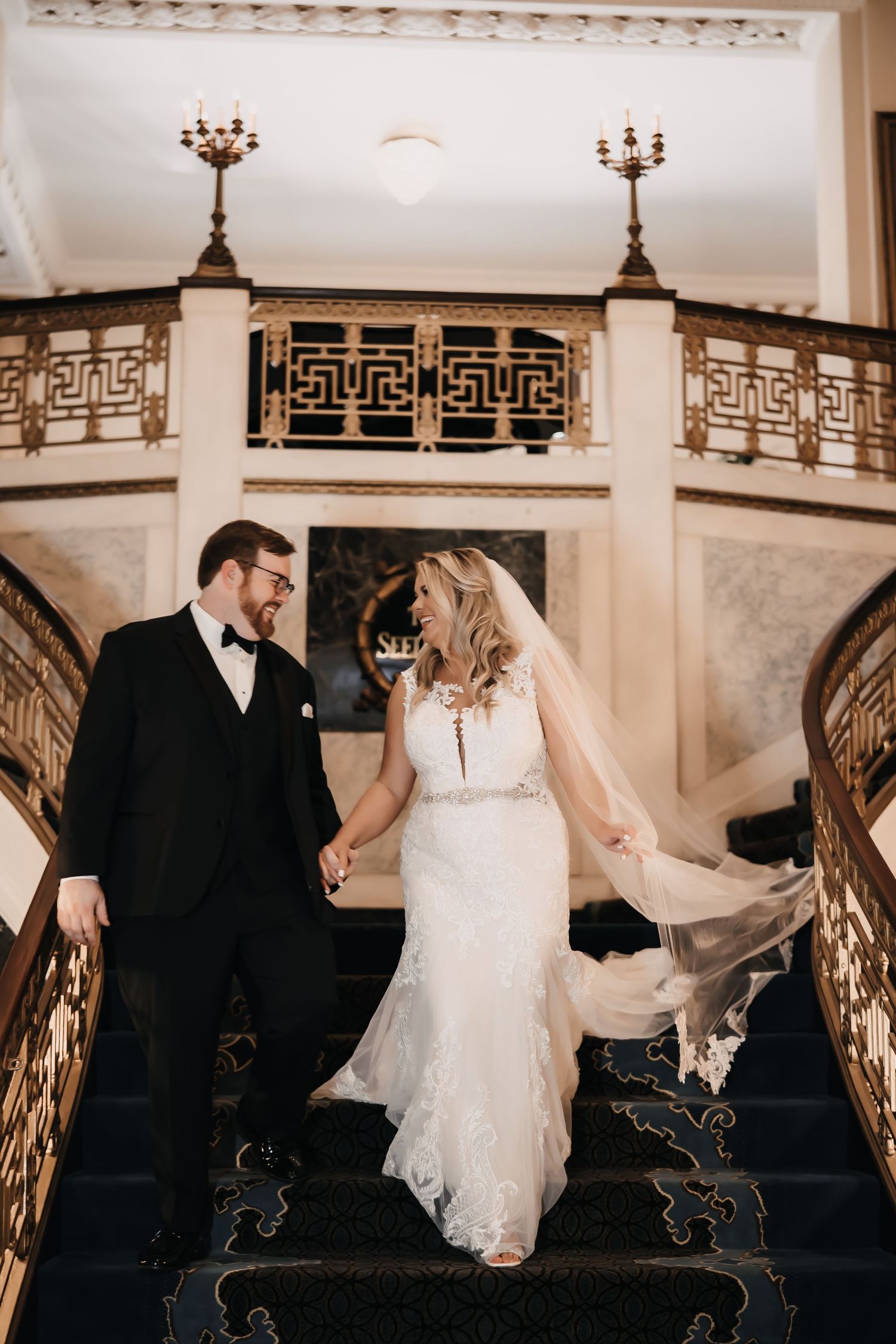 A couple in wedding attire descends an elegant staircase with ornate railings and vintage candelabras.