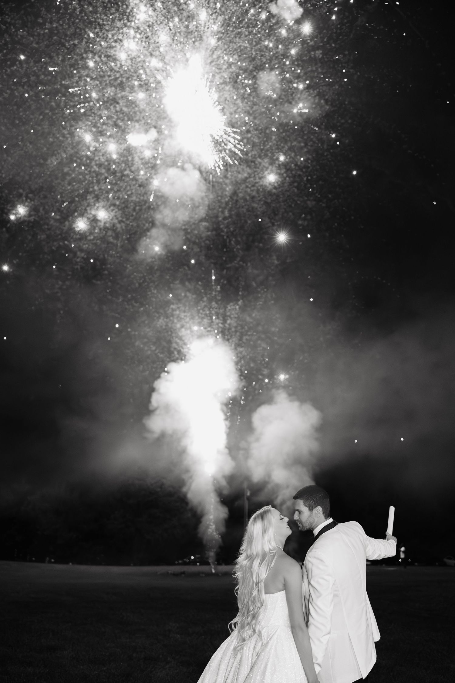 Bride and groom kiss under spectacular fireworks display in romantic black and white wedding photo.