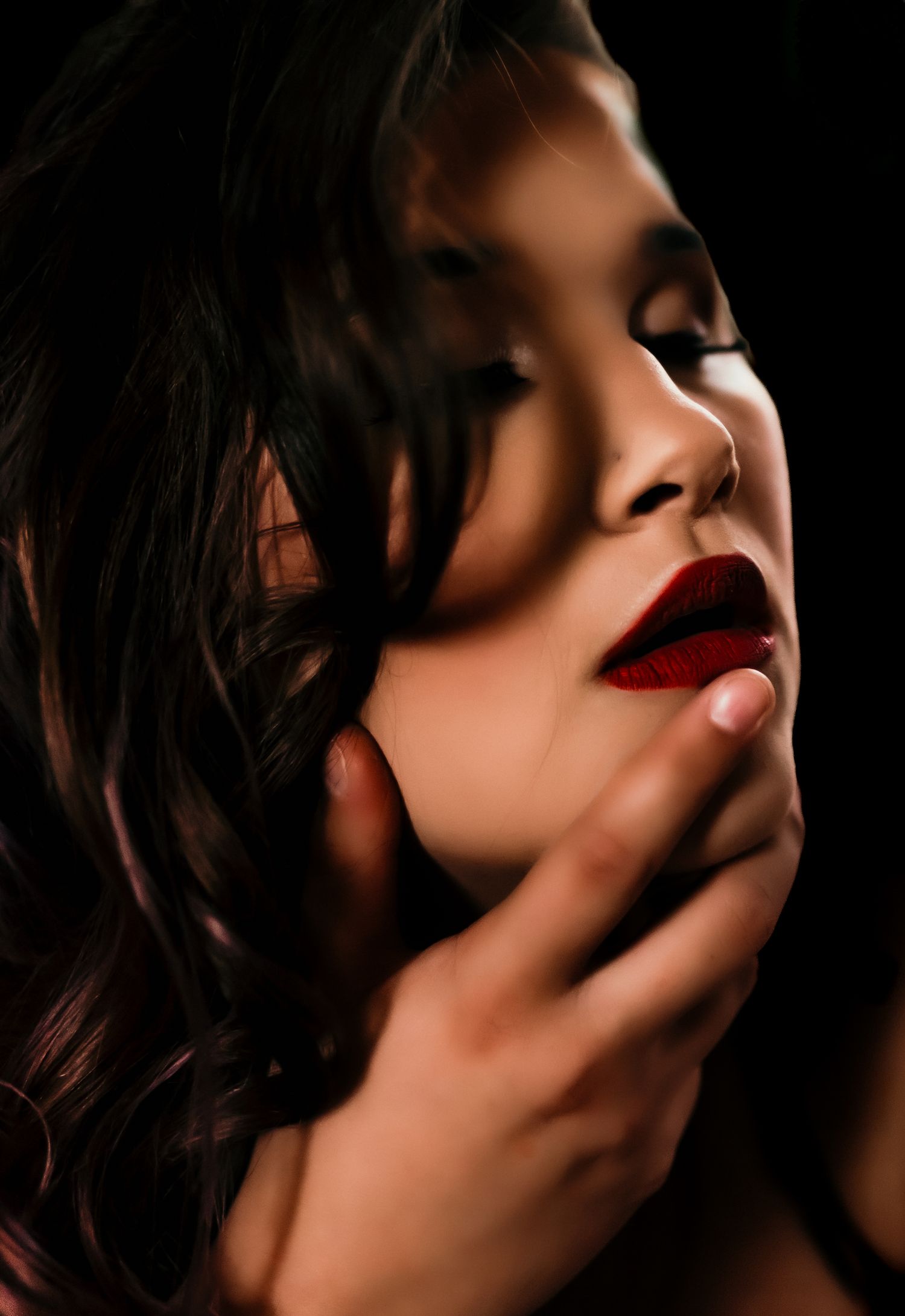 A woman with red lips and dark hair poses dramatically in low-key lighting with a hand near her chin.
