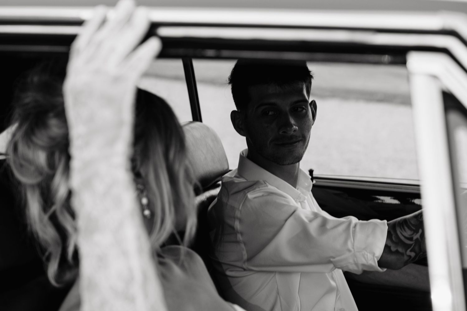 Black and white wedding photo of groom in white shirt looking back from vintage vehicle at blonde bride in lace dress.
