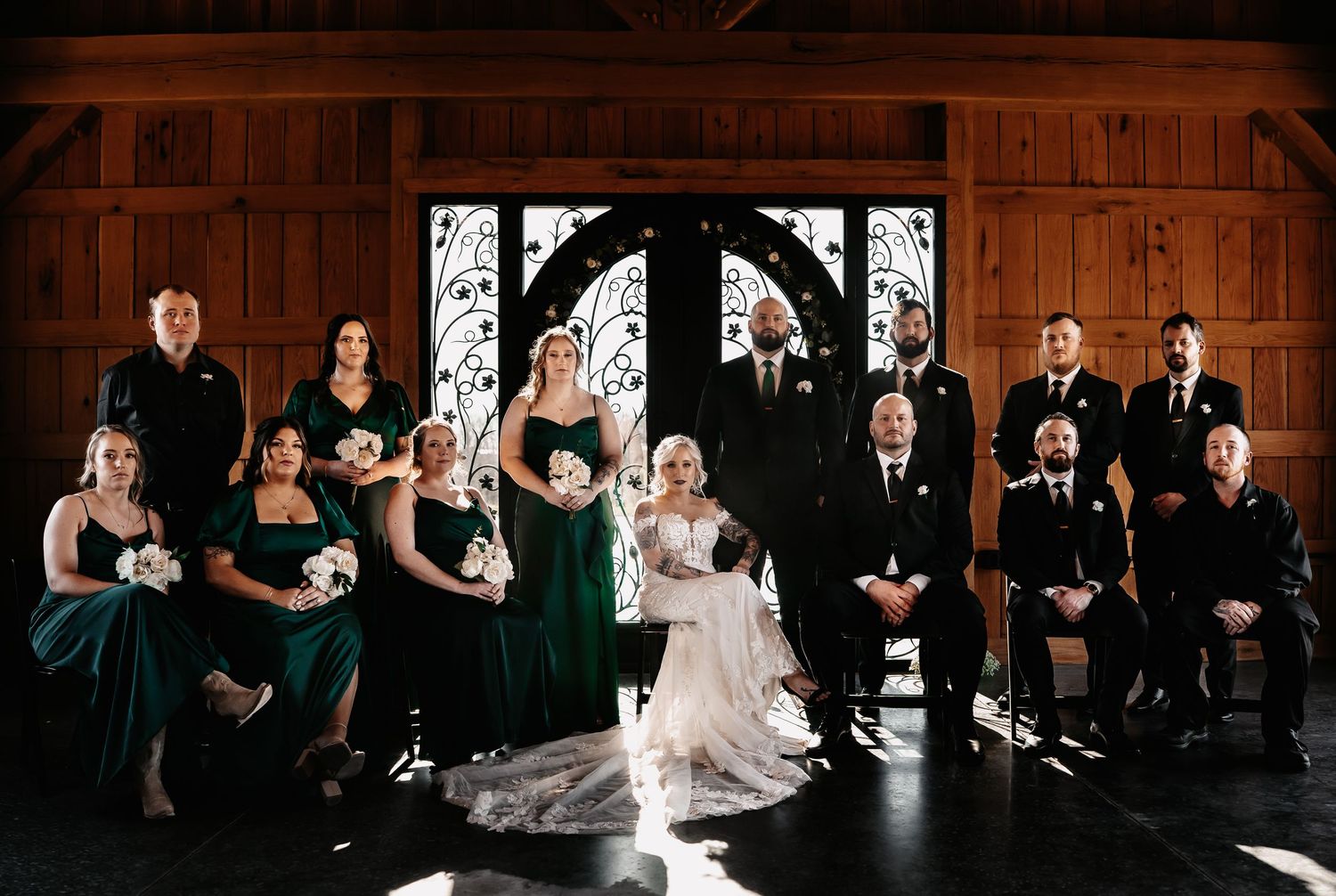 Wedding party in green dresses and black suits posed before ornate doors in a rustic wood-paneled venue.