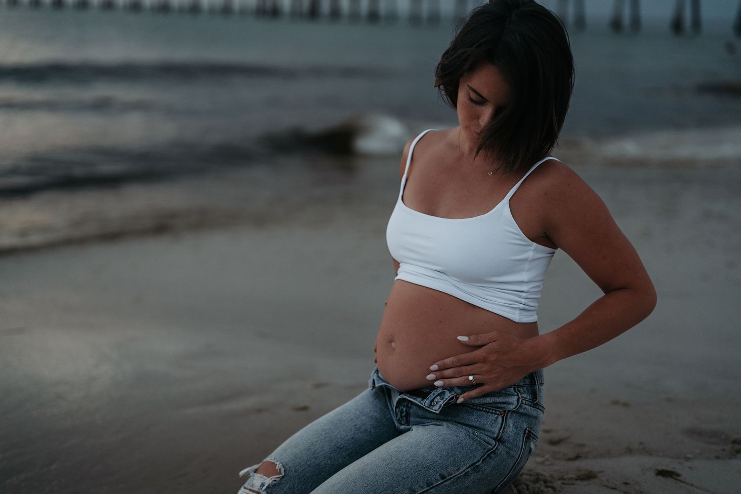 Pregnant woman in white crop top and jeans sitting on beach, cradling baby bump near ocean waves.