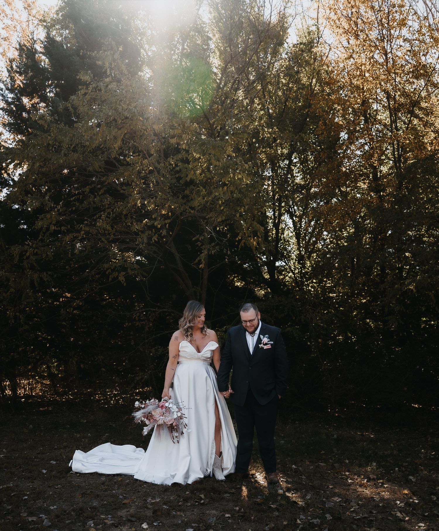 A couple in formal wedding attire walk together through an autumn forest setting with golden leaves and natural light.