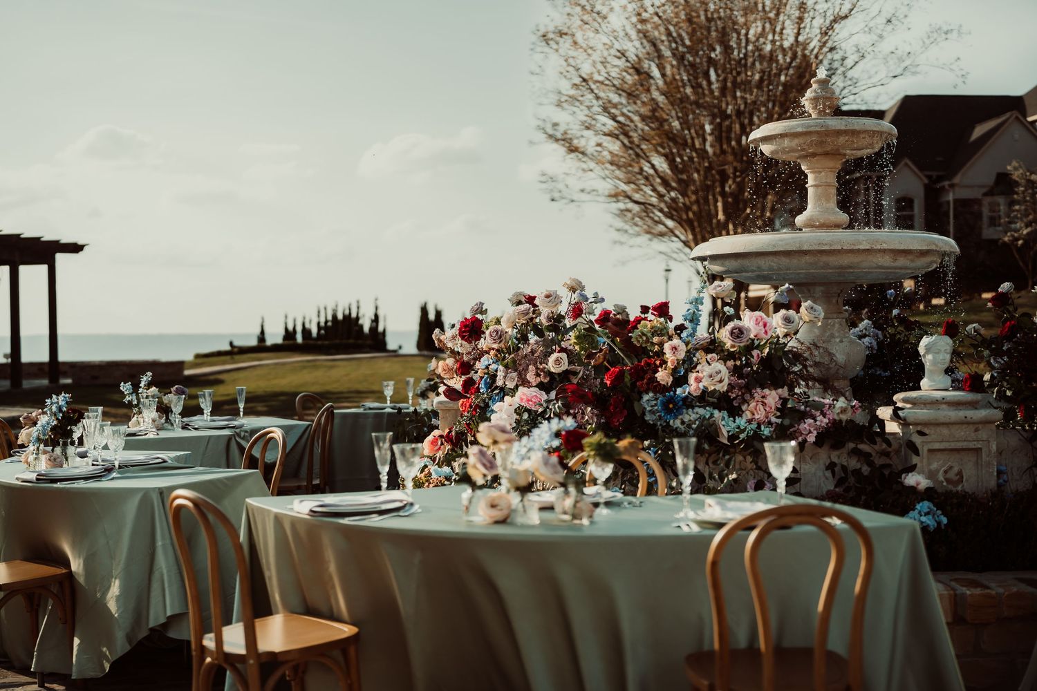 Elegant outdoor wedding reception table with sage linens, lush floral centerpiece, and stone fountain near waterfront venue.