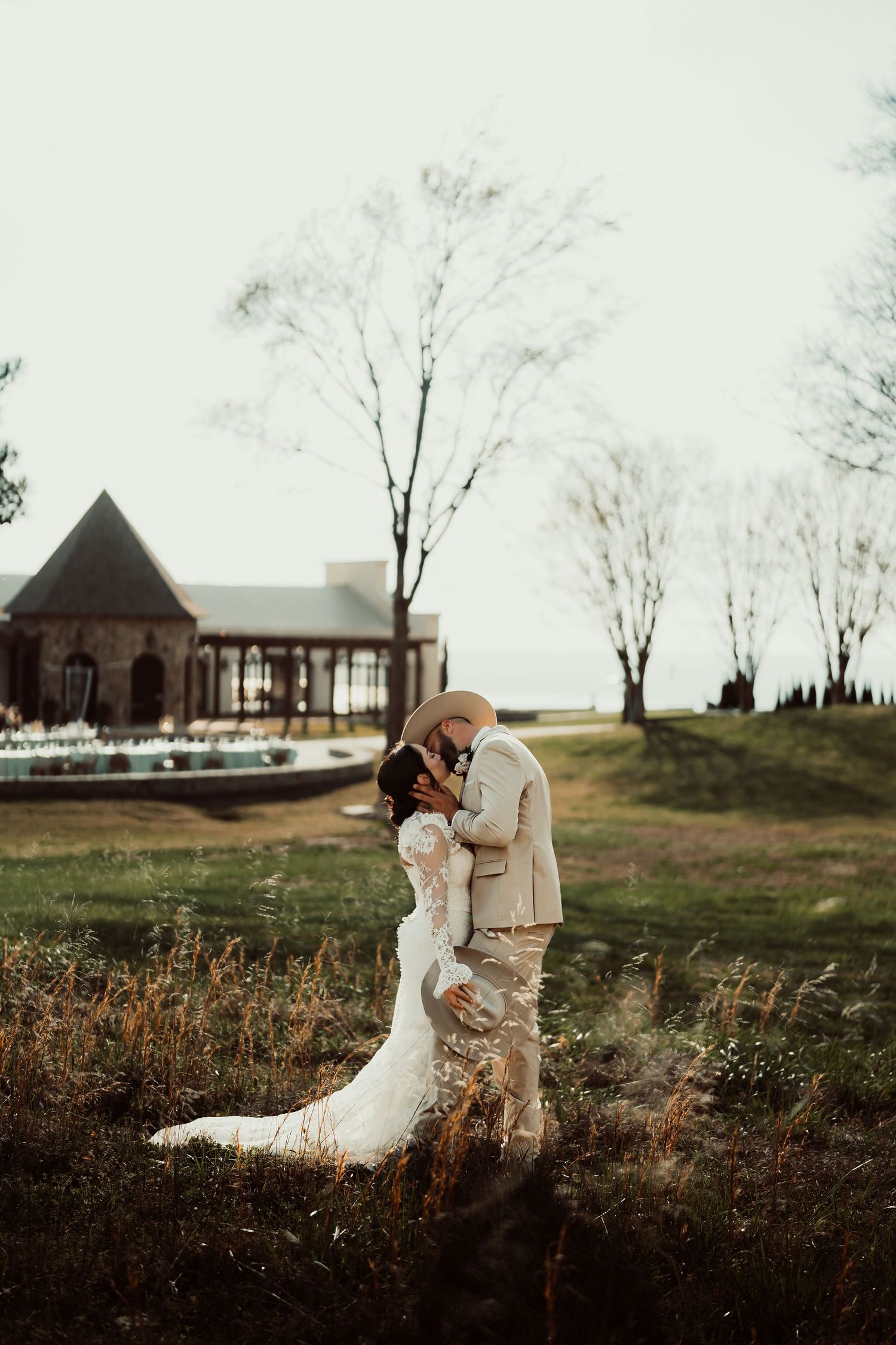 Bride and groom share a romantic kiss in a lace wedding gown and tan suit with cowboy hat outdoors.